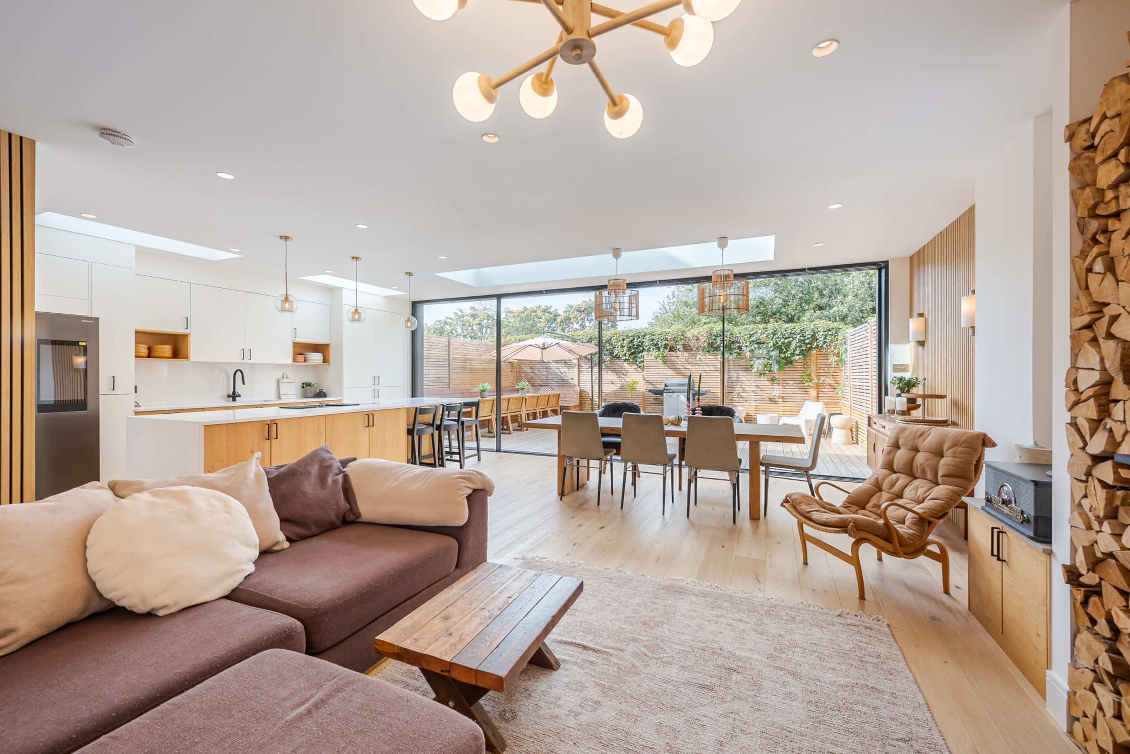 A modern living and dining area with large windows, featuring a kitchen on one side, a dining table in the center, and a cozy seating arrangement with a rug in front.