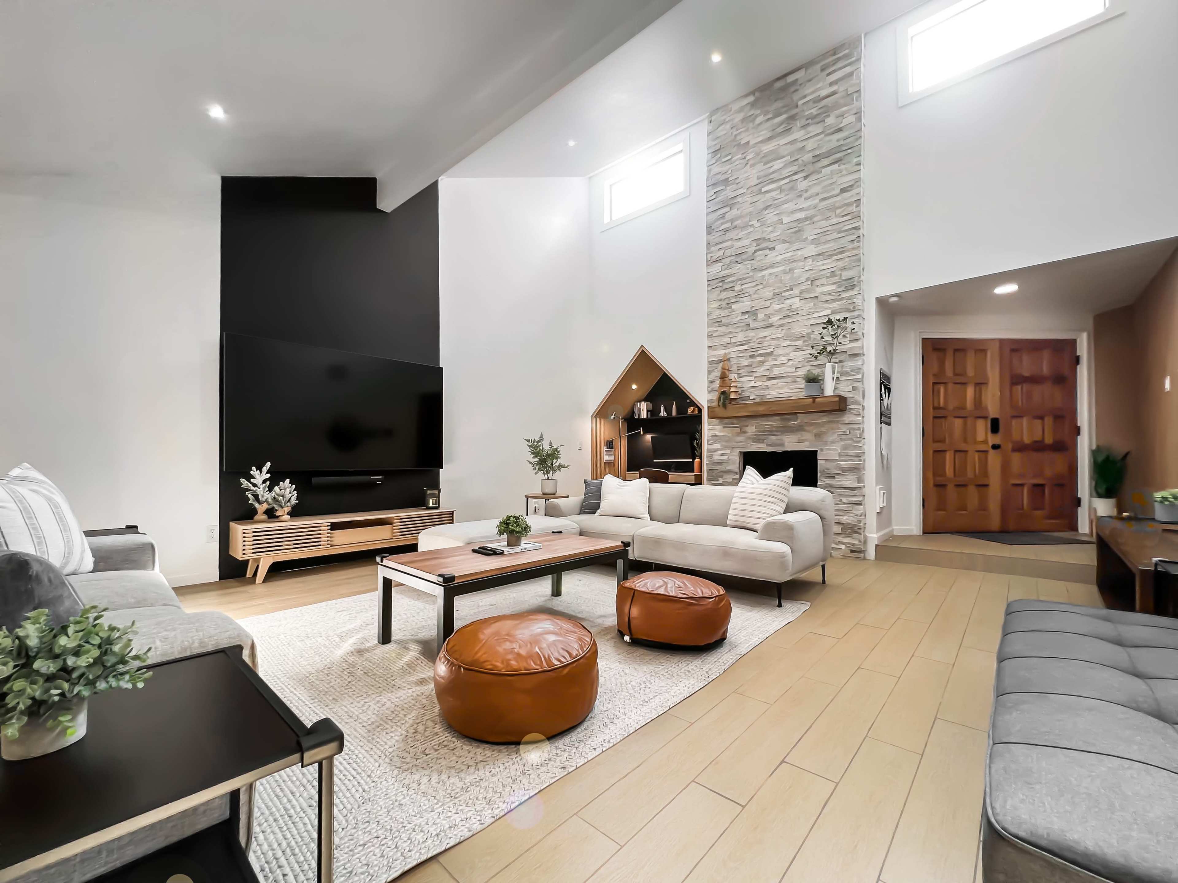A modern living room featuring a large flat-screen TV, a stone fireplace, and a mix of seating including a sectional sofa and leather ottomans.