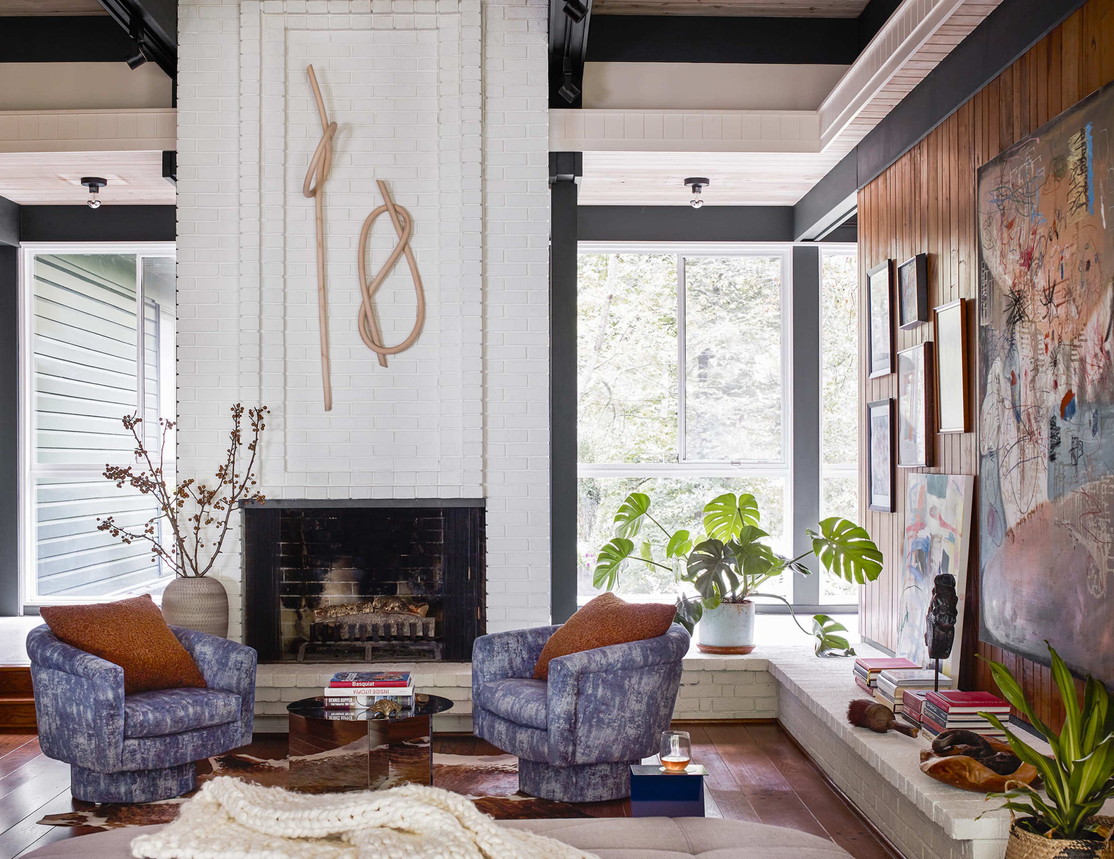 The image shows a living room with two patterned blue chairs facing a white brick fireplace, surrounded by large windows and plants.