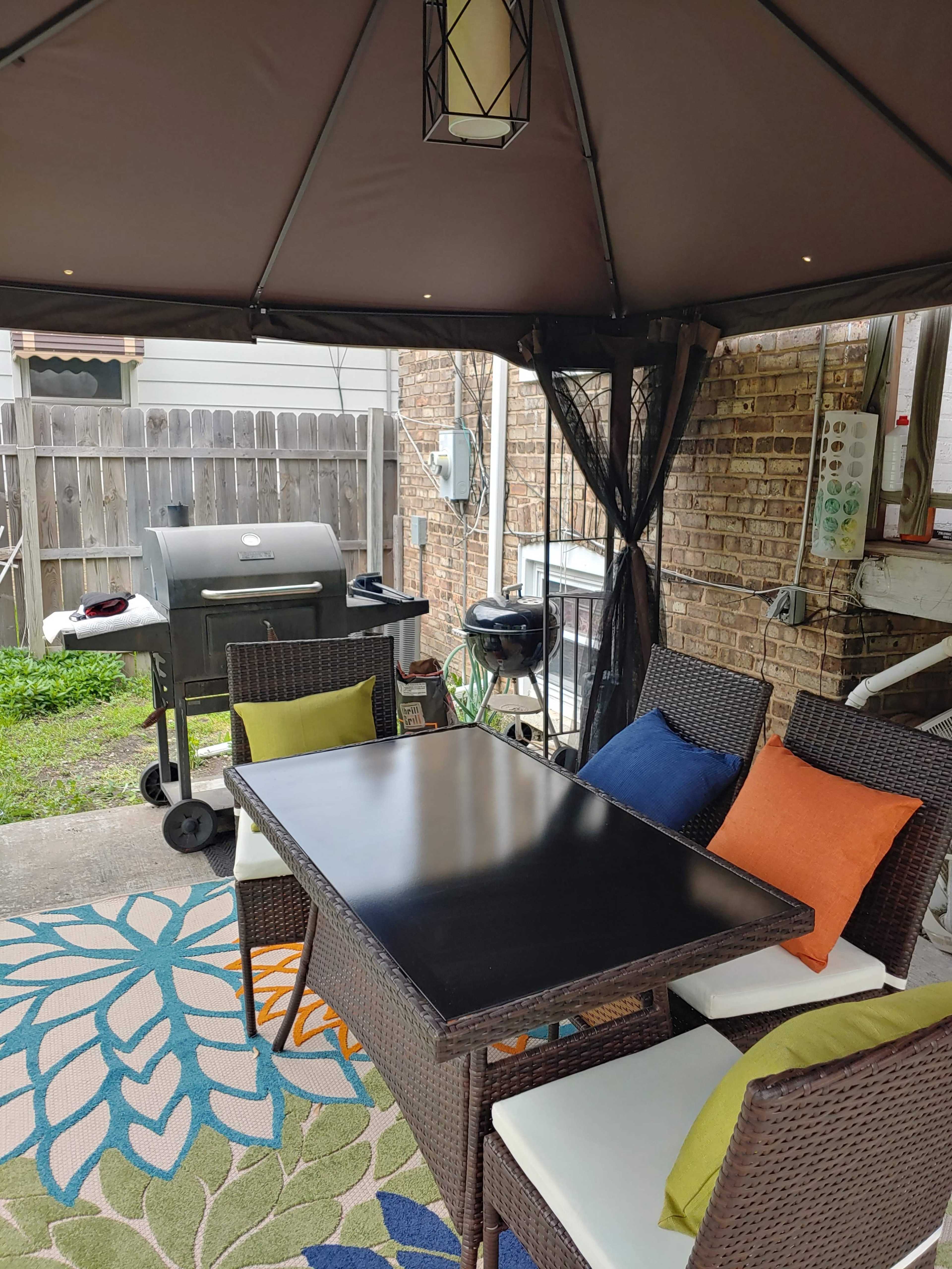 A patio area features a black table surrounded by four chairs, with a grill positioned nearby under a gazebo.
