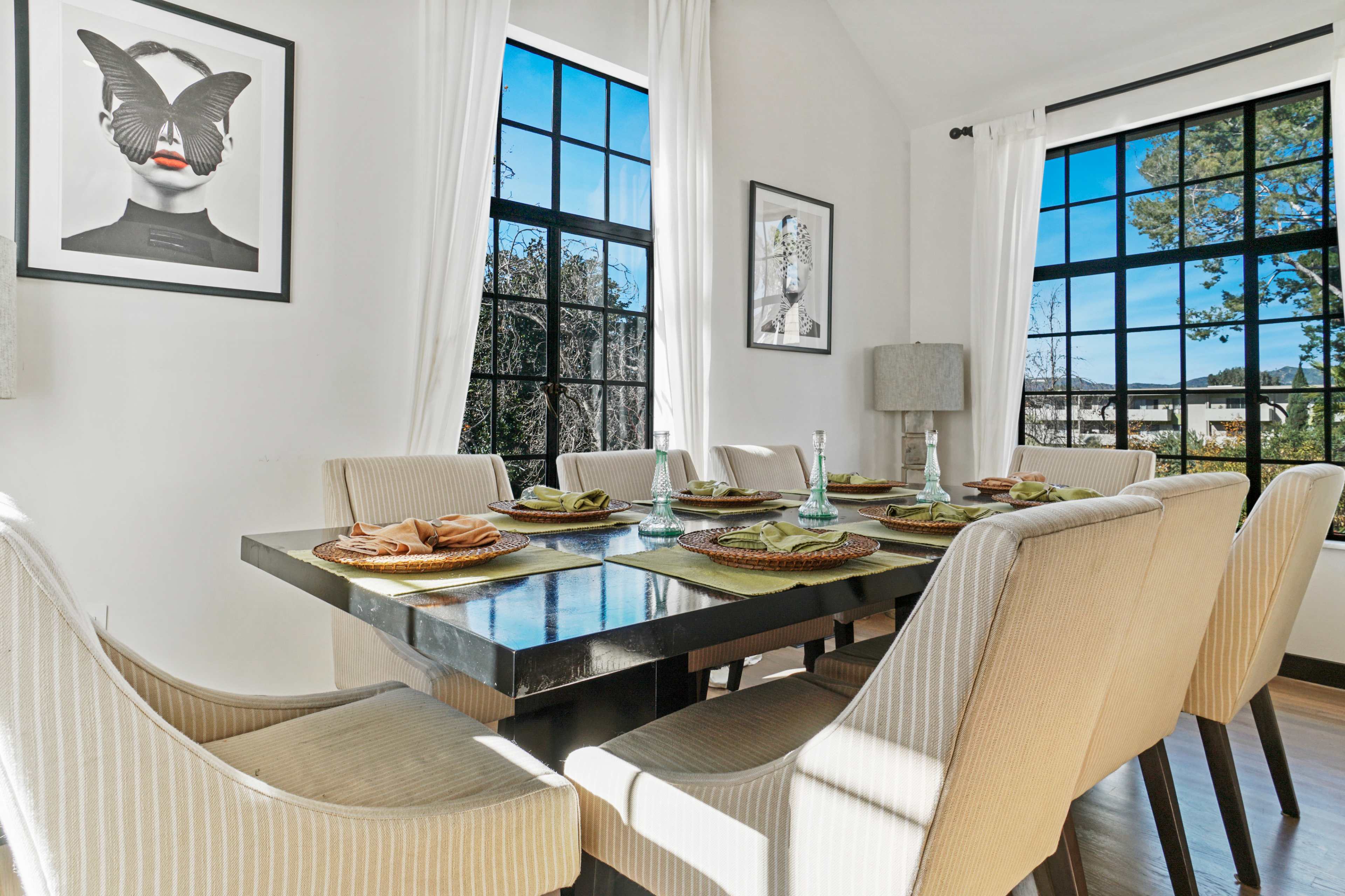 A dining room features a large table set for eight with a modern design, surrounded by beige upholstered chairs, and large windows letting in natural light.