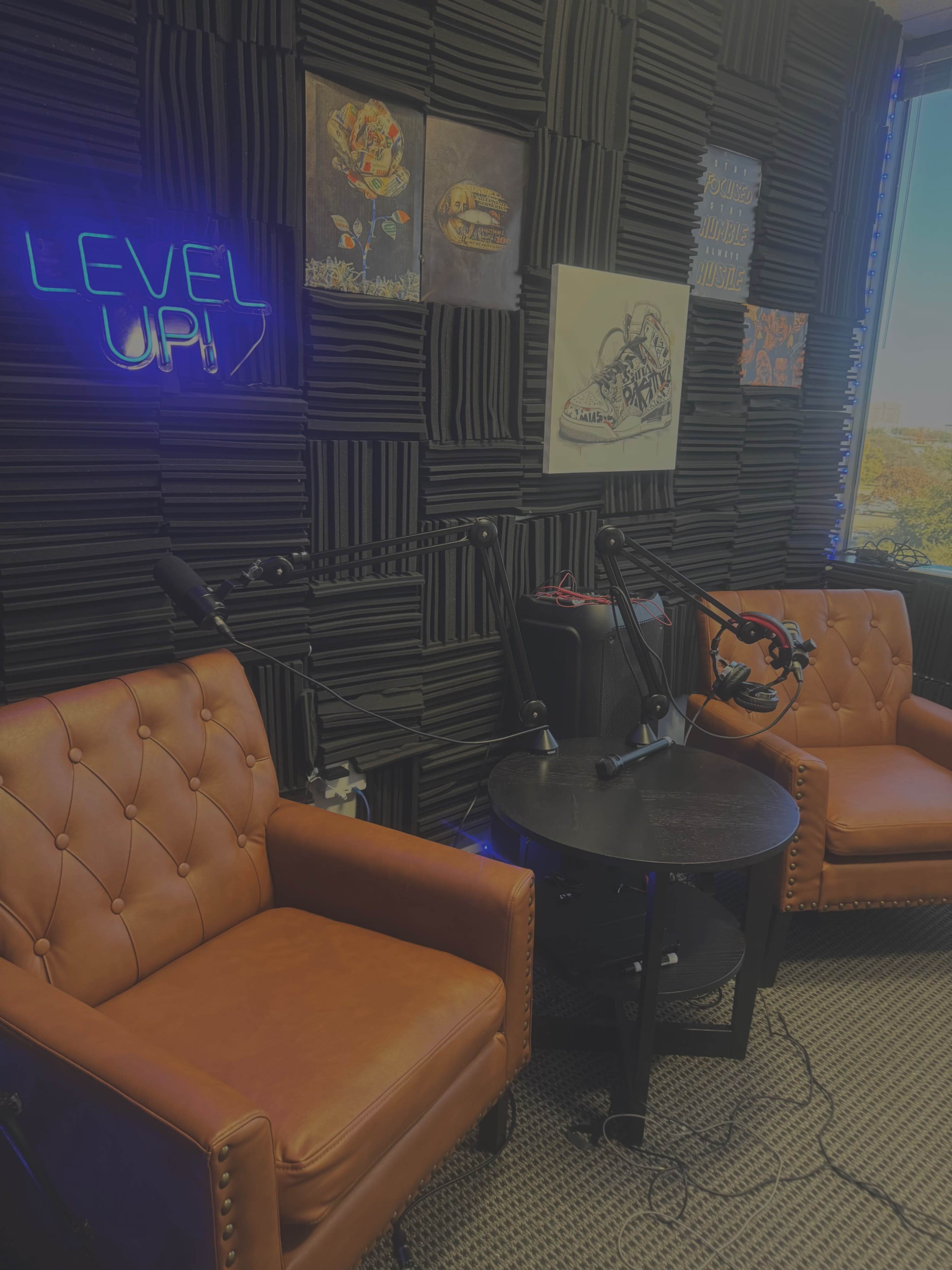 The image shows a podcast studio featuring two brown leather chairs, a small round table, and soundproof walls adorned with various posters, alongside a neon sign that reads "LEVEL UP."