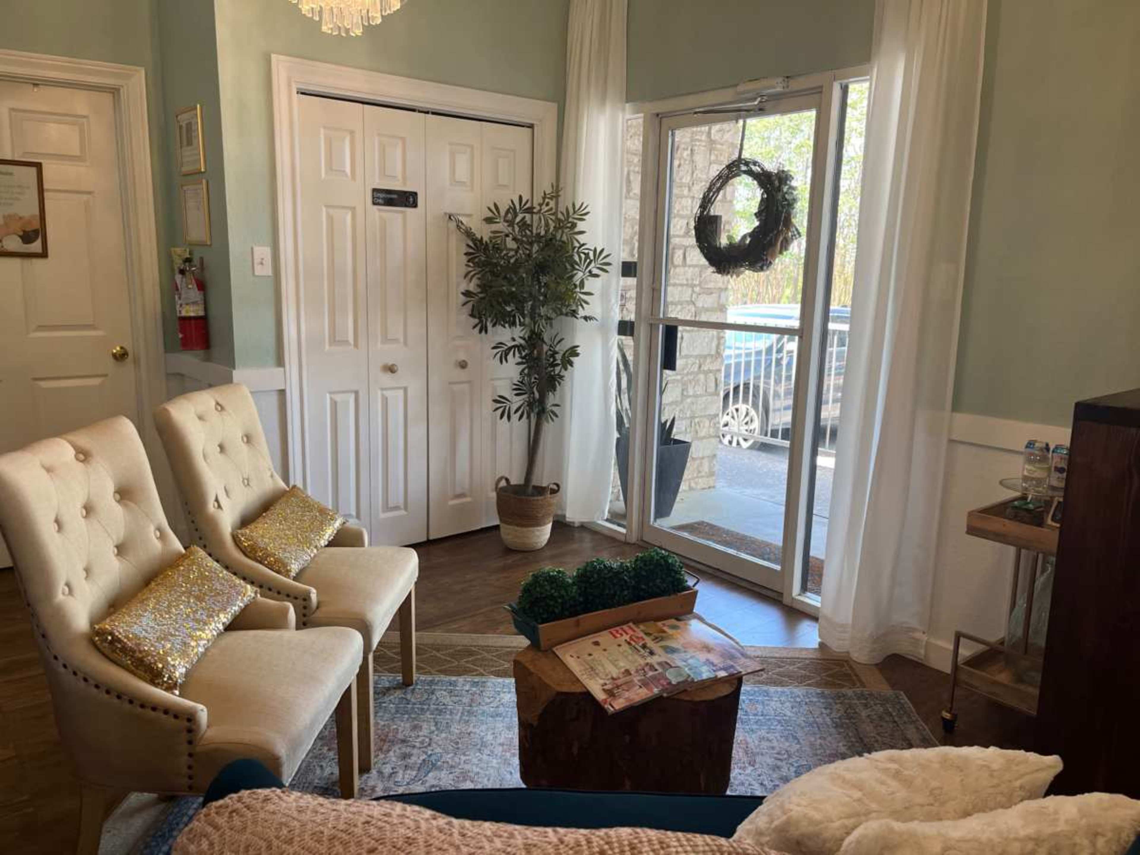 The image shows a cozy waiting area with two beige chairs, a small wooden table, and a glass door leading outside, surrounded by decorative plants and a plush rug.