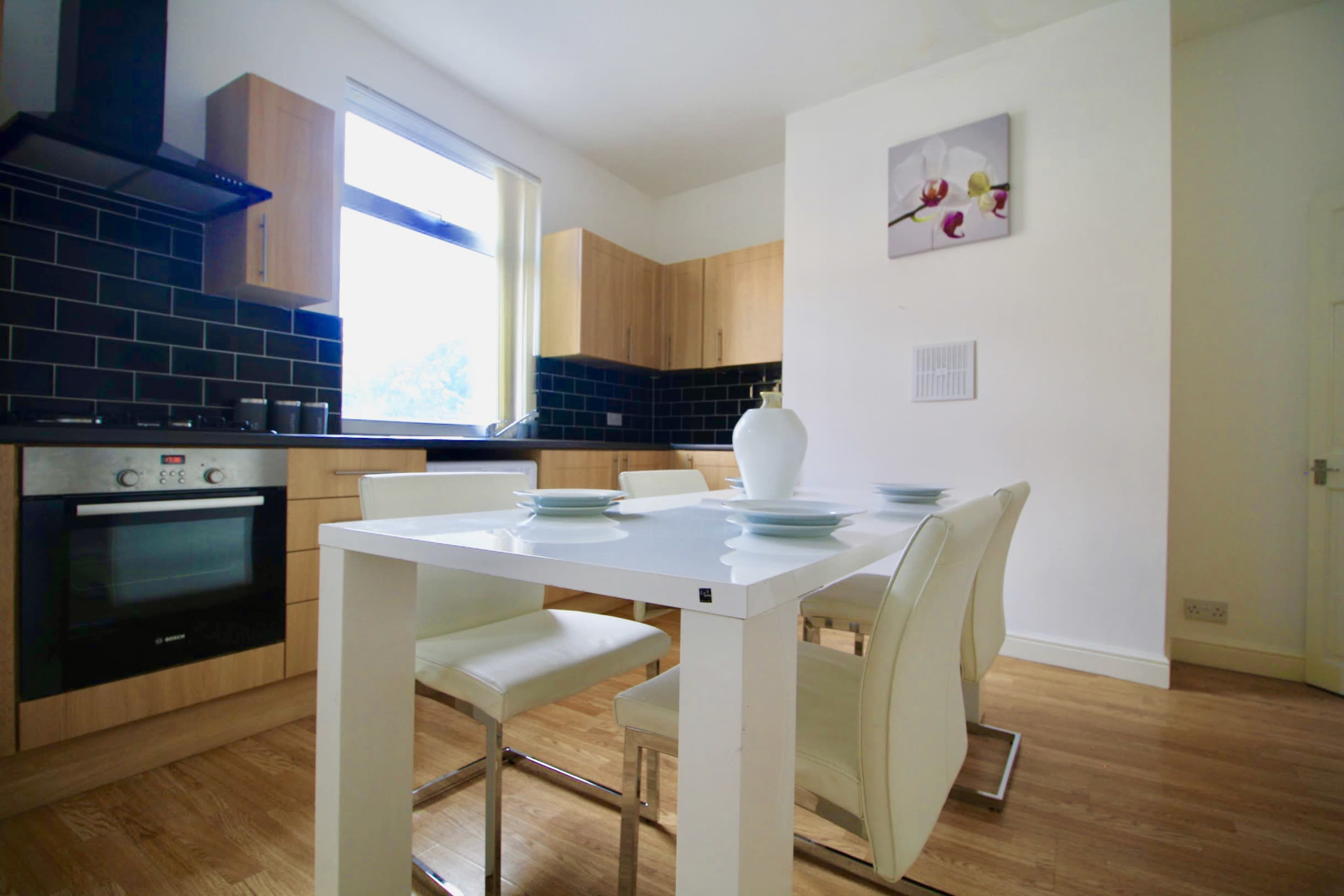A modern kitchen features wooden cabinetry, black tiled walls, and a white dining table with four chairs.