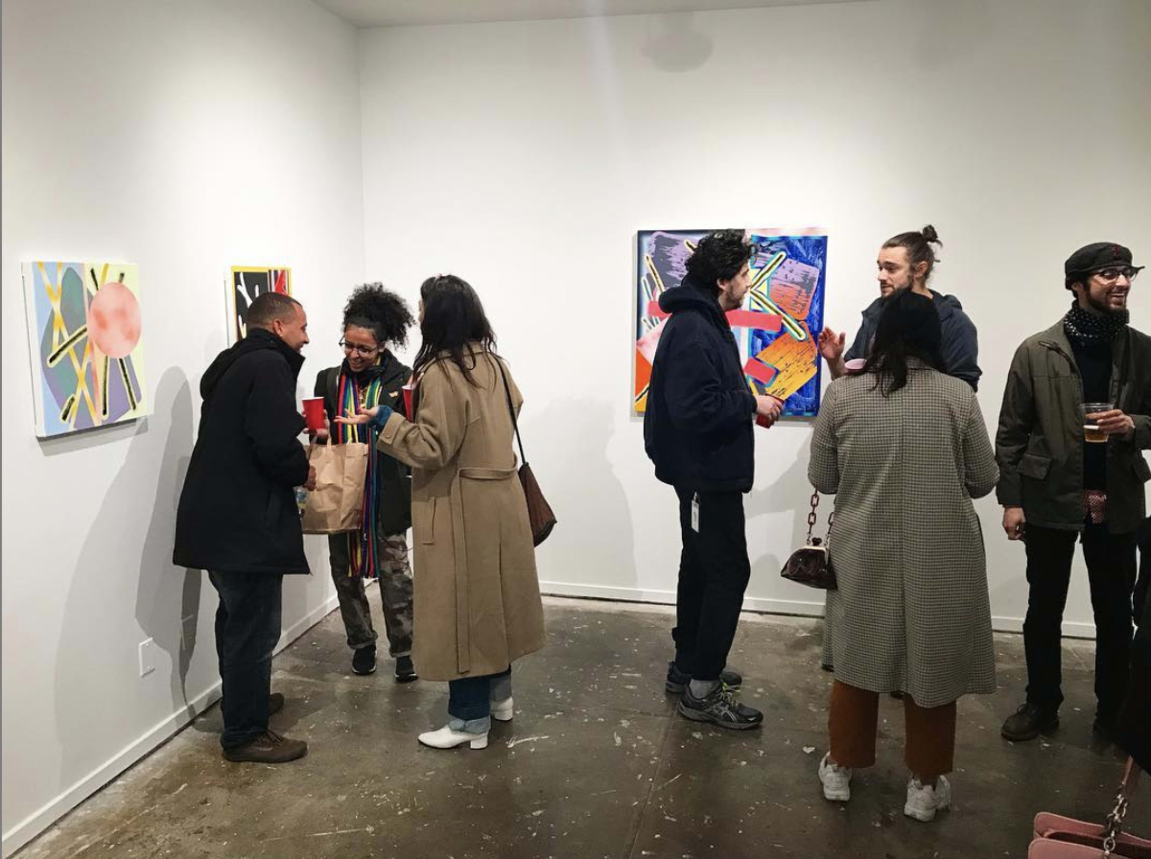 A group of people gathers in an art gallery, engaging in conversation while observing colorful artwork on the walls.