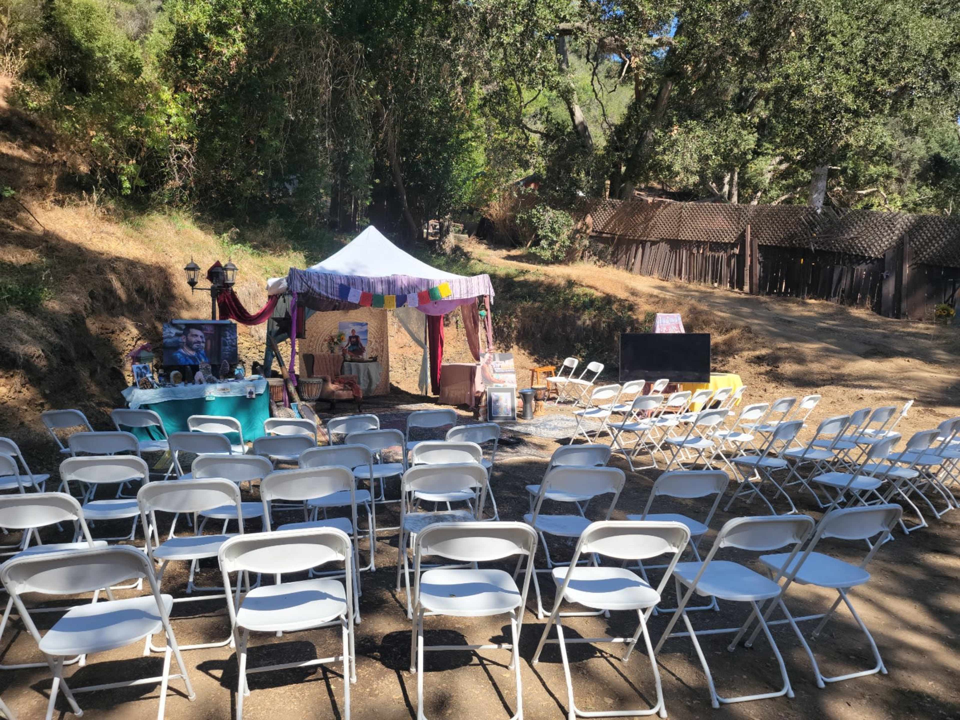 A tent with colorful decorations is set up in a clearing, facing rows of white folding chairs, with a rustic building visible in the background.