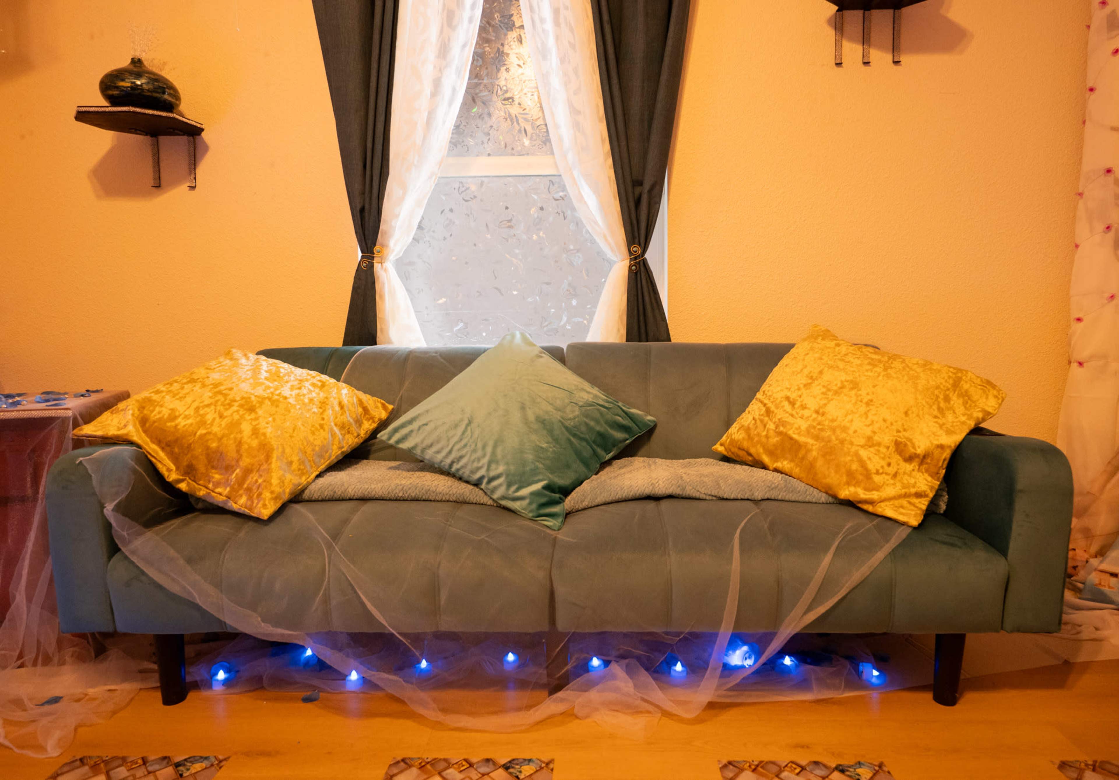 A turquoise sofa adorned with gold and teal cushions is positioned near a window, draped with sheer curtains and illuminated by blue lights underneath.