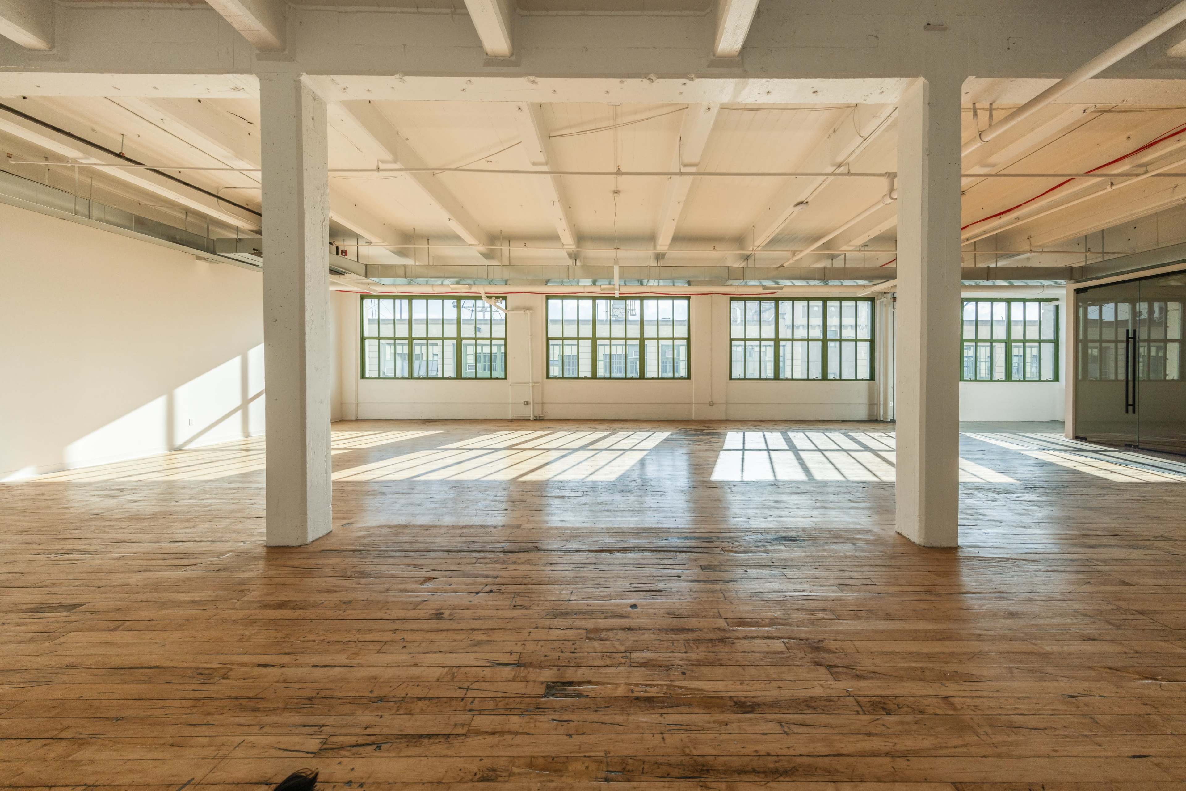 The image depicts a spacious, empty room with wooden floors and large windows allowing natural light to illuminate the space.