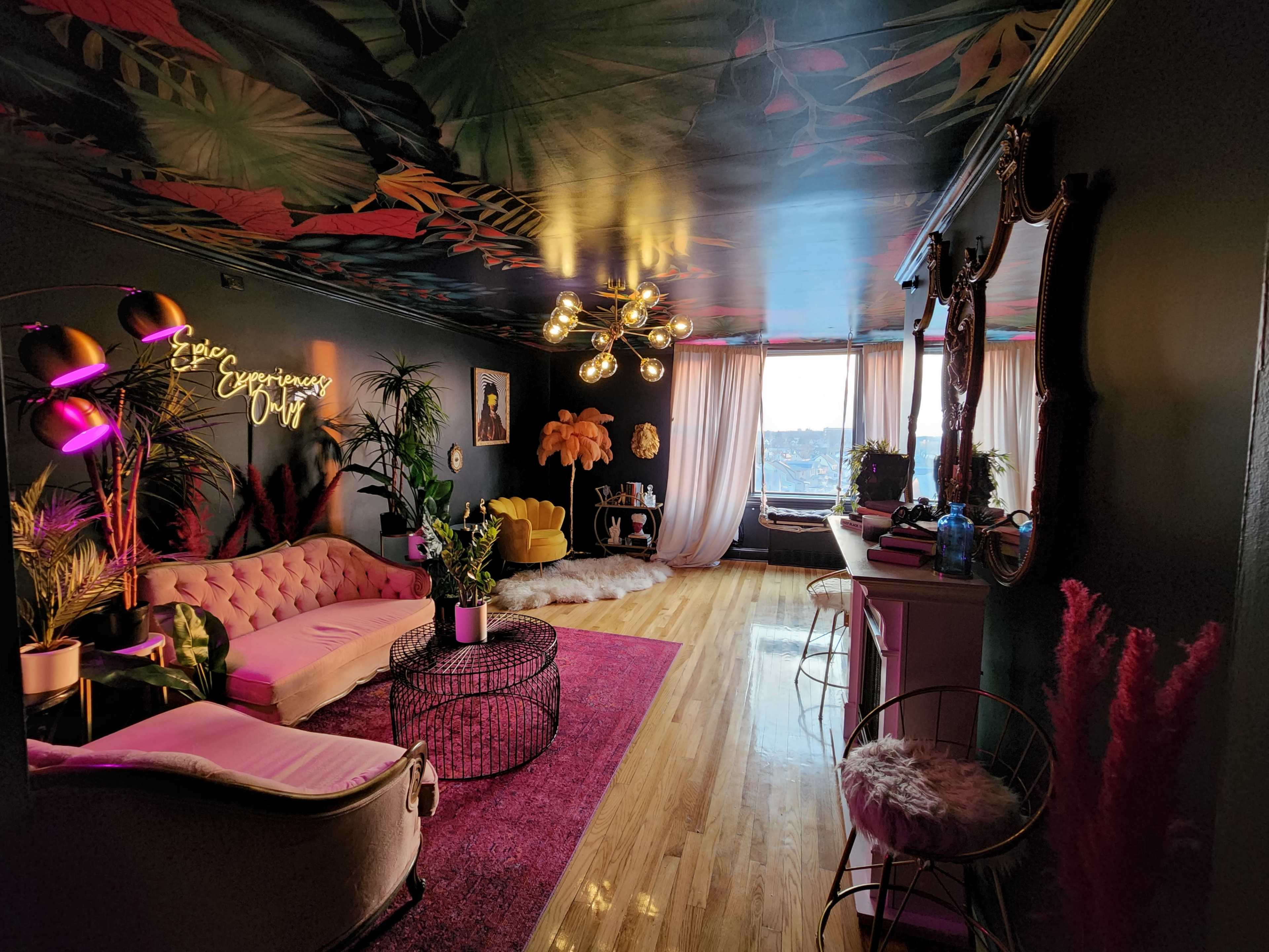 A colorful living room with a pink velvet sofa, a round black coffee table, tropical plant decor, and large windows providing natural light.