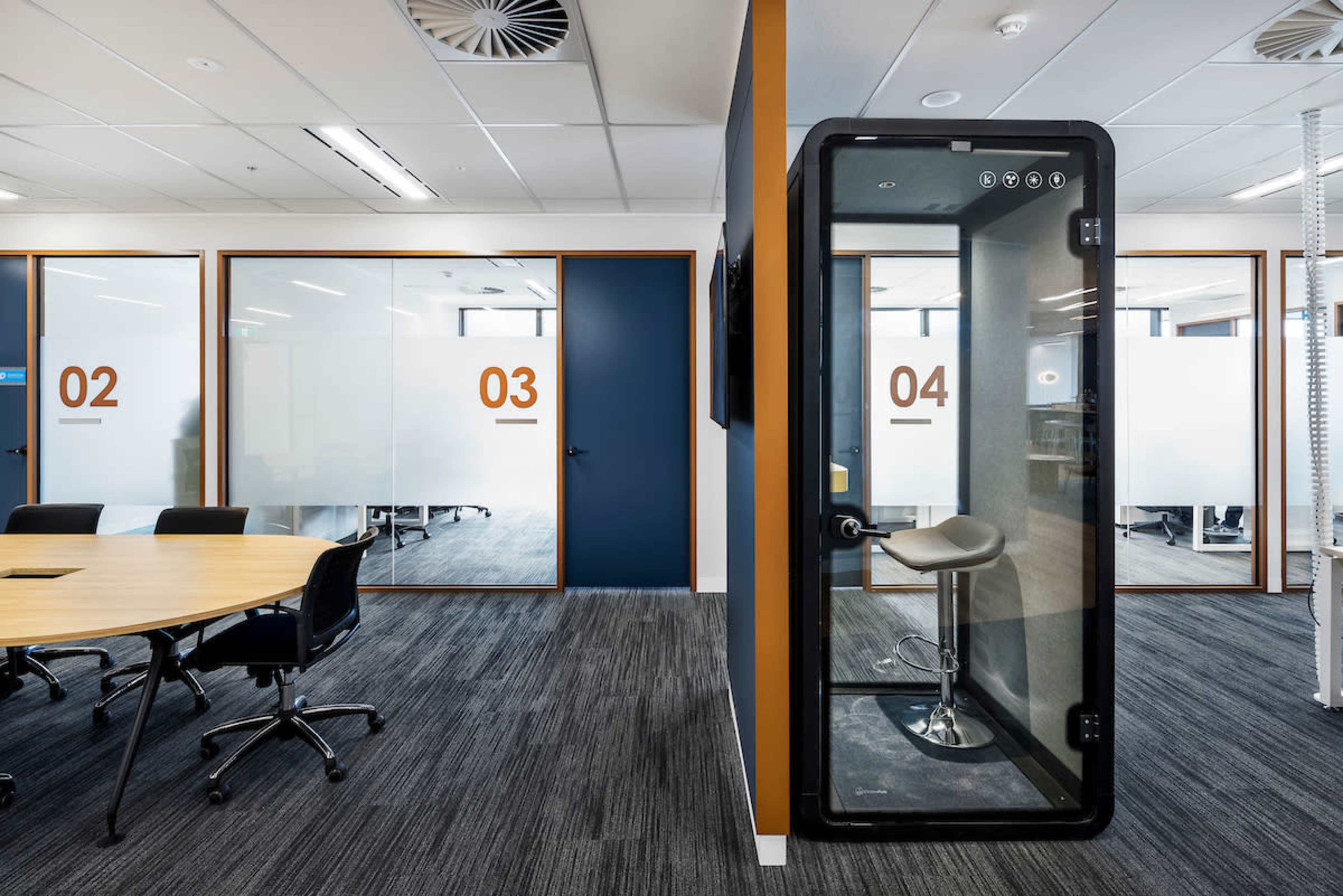 The image shows a modern office space featuring glass meeting rooms numbered 02, 03, and 04, with a round conference table and a private phone booth.