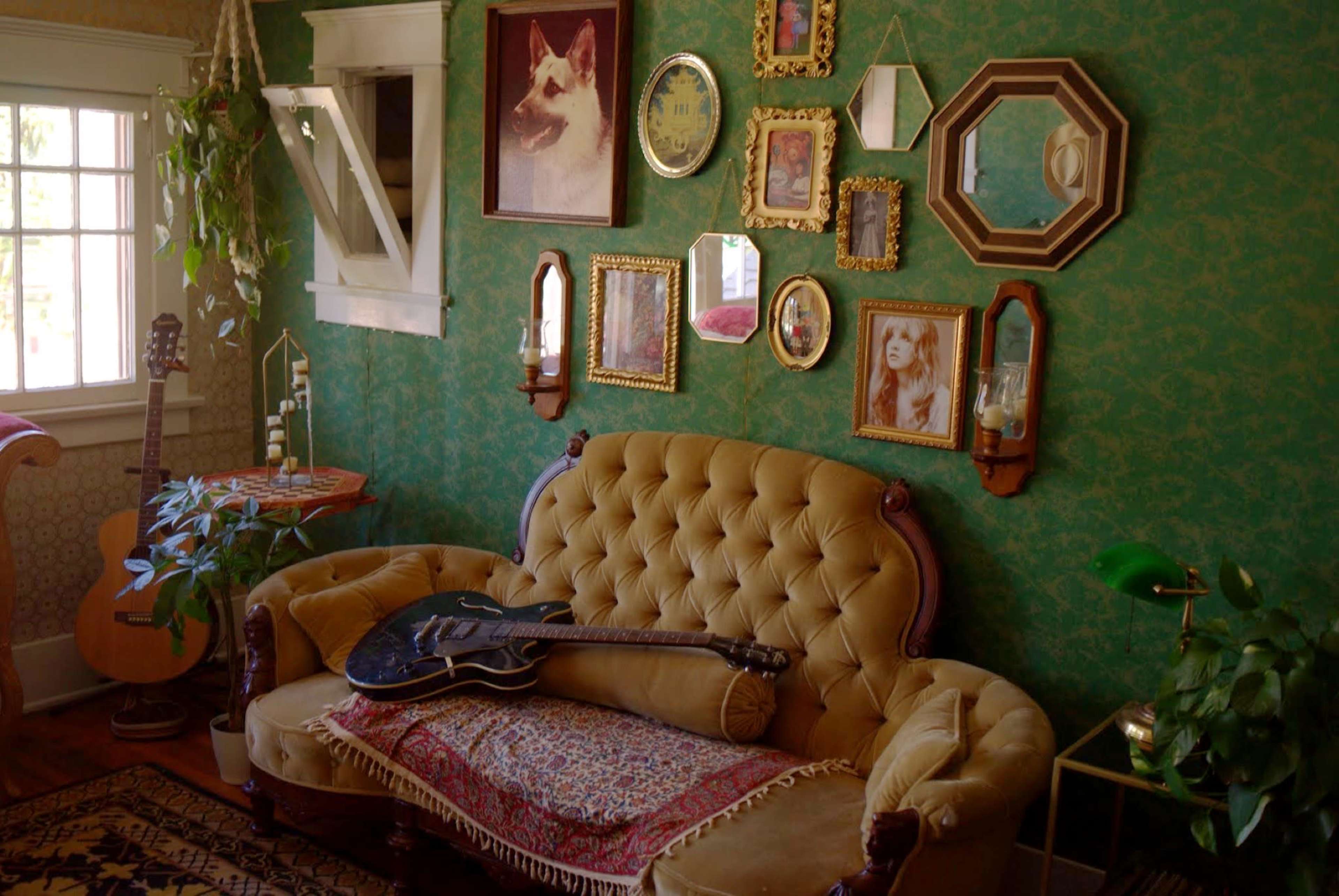 A vintage living room with a green patterned wallpaper, a tufted yellow sofa adorned with a woven blanket, and an array of framed pictures on the wall.