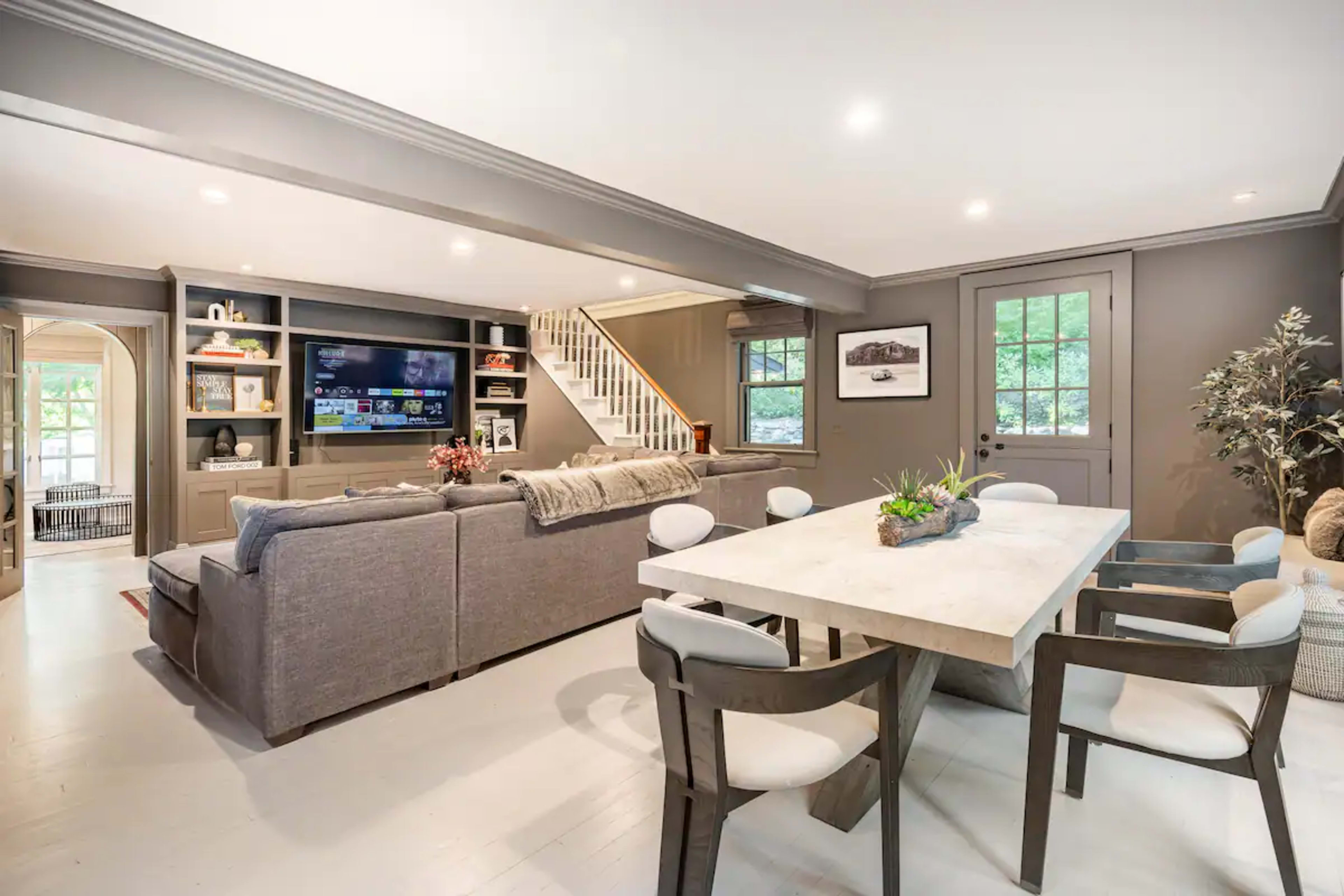 A spacious living area features a gray sofa, a large television on a built-in shelf, and a dining table surrounded by modern chairs.