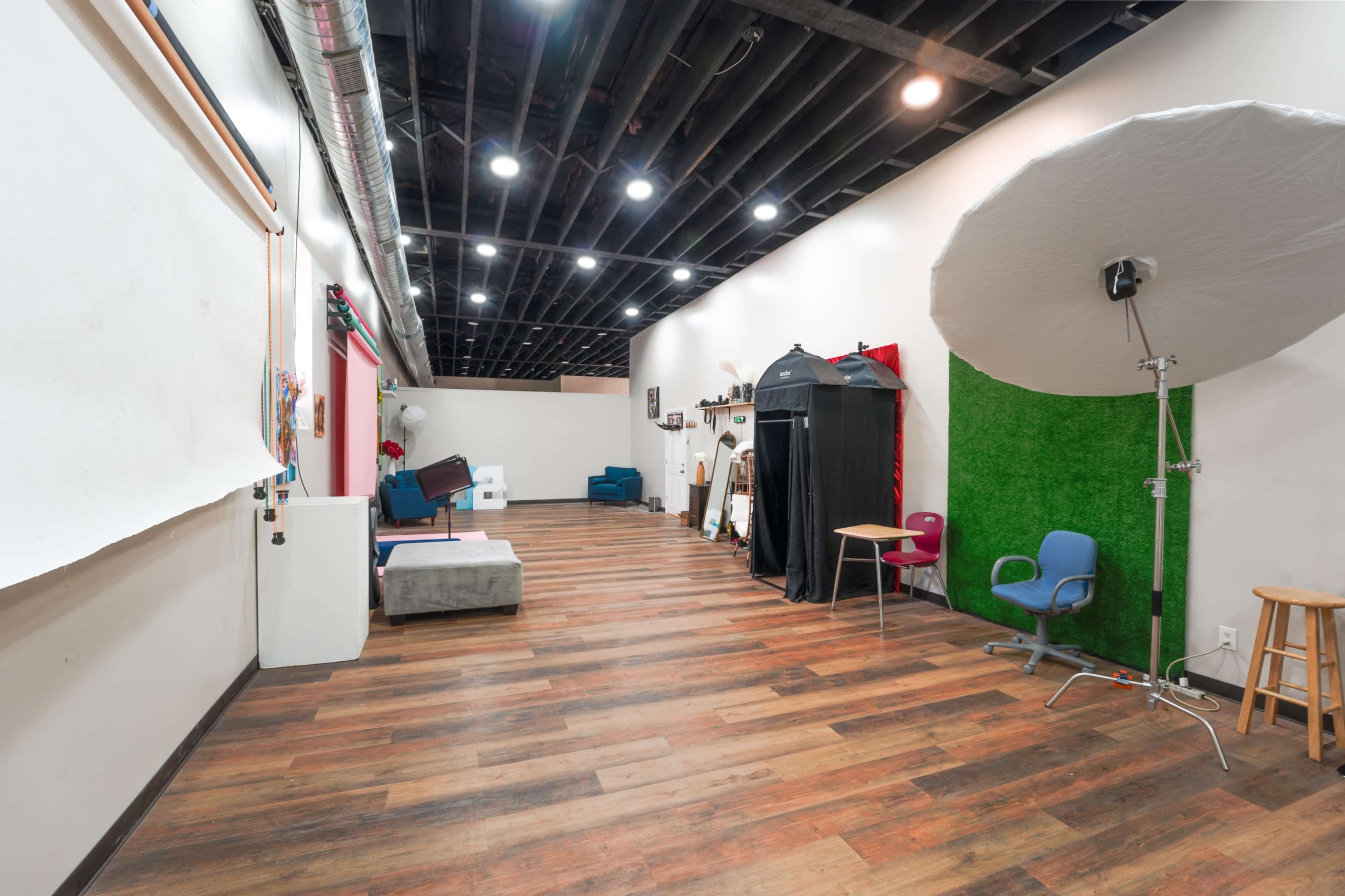 The image shows a spacious studio with wooden flooring, brightly lit by overhead lights, featuring various props and a backdrop setup along one wall.
