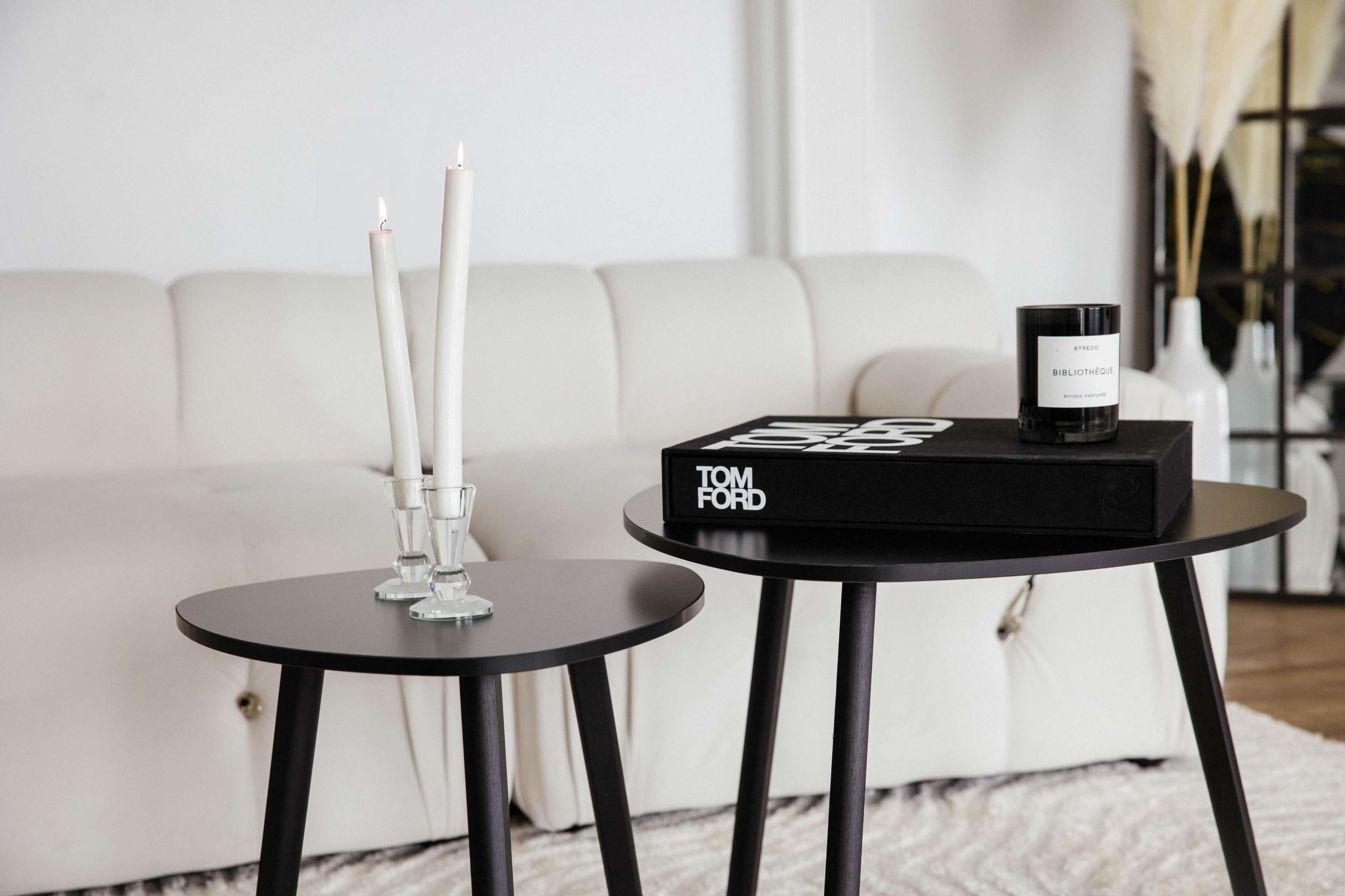 A black side table holds a Tom Ford box and a candle, while a second small table features two white candles in glass holders, all set against a white sofa.