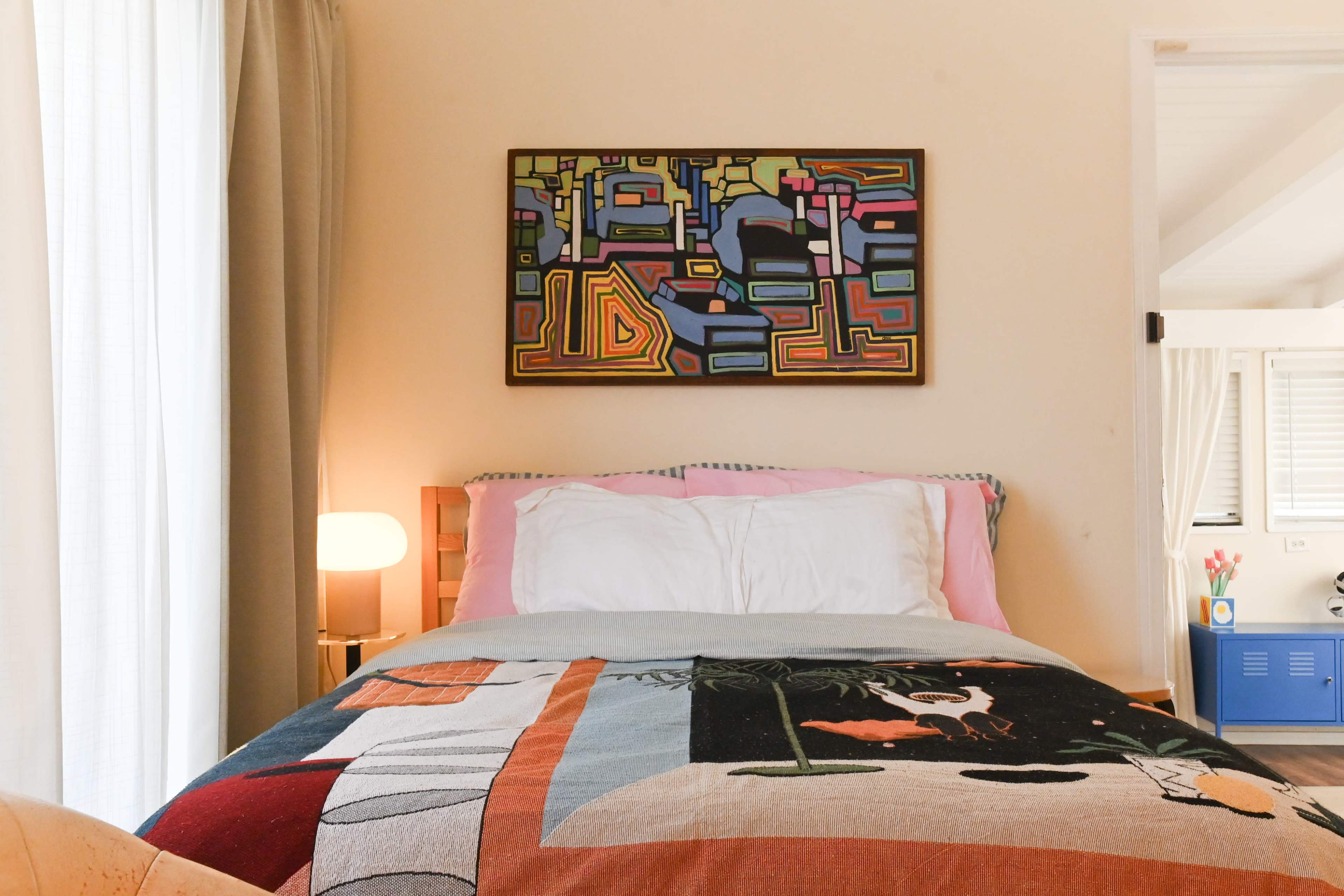 A bed with colorful bedding is positioned in a well-lit room featuring a large painting above it and a lamp beside the bed.
