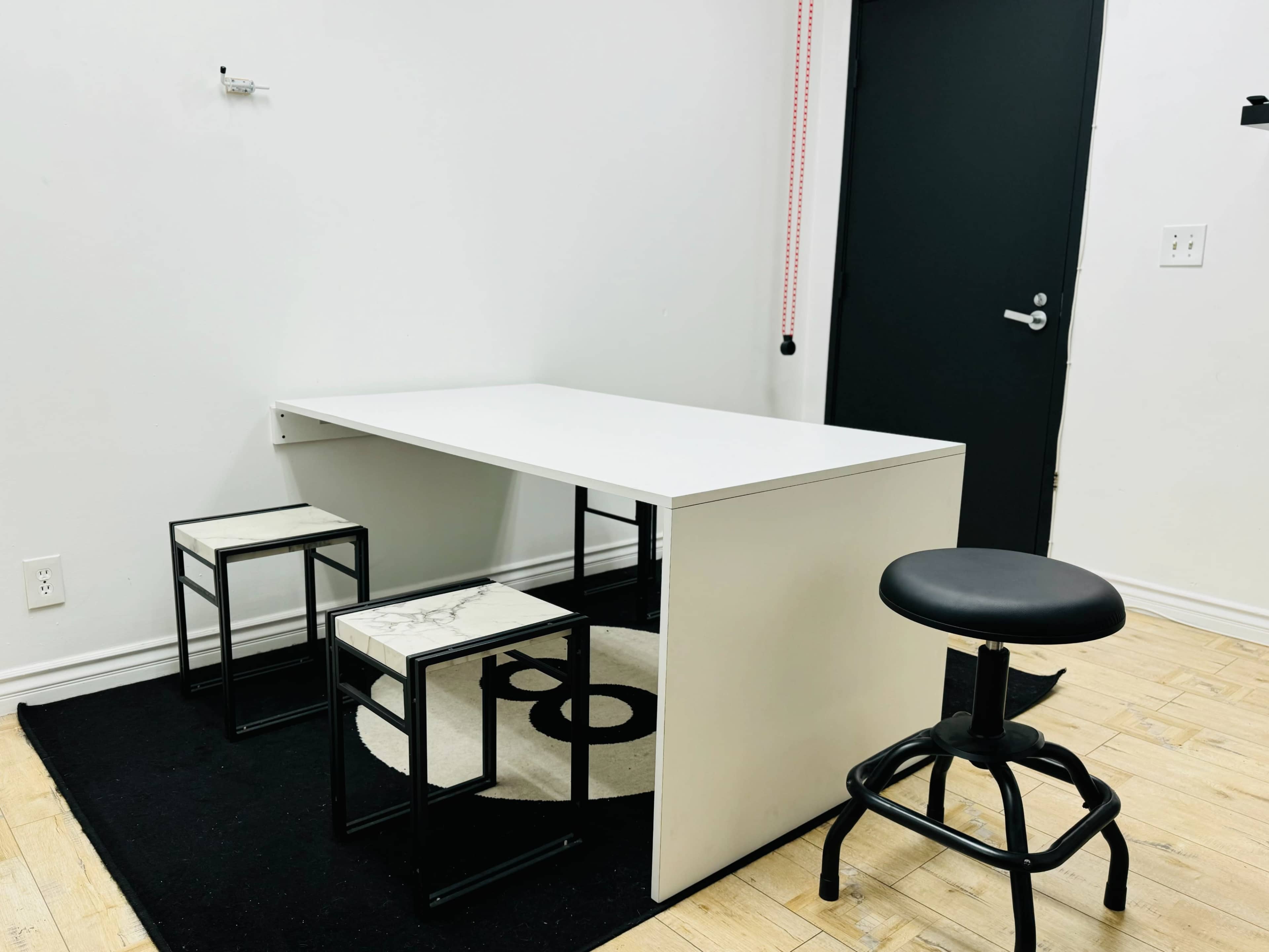 A minimalist workspace features a white table with two small stools and a black swivel stool, all placed on a patterned black rug against a plain white wall.