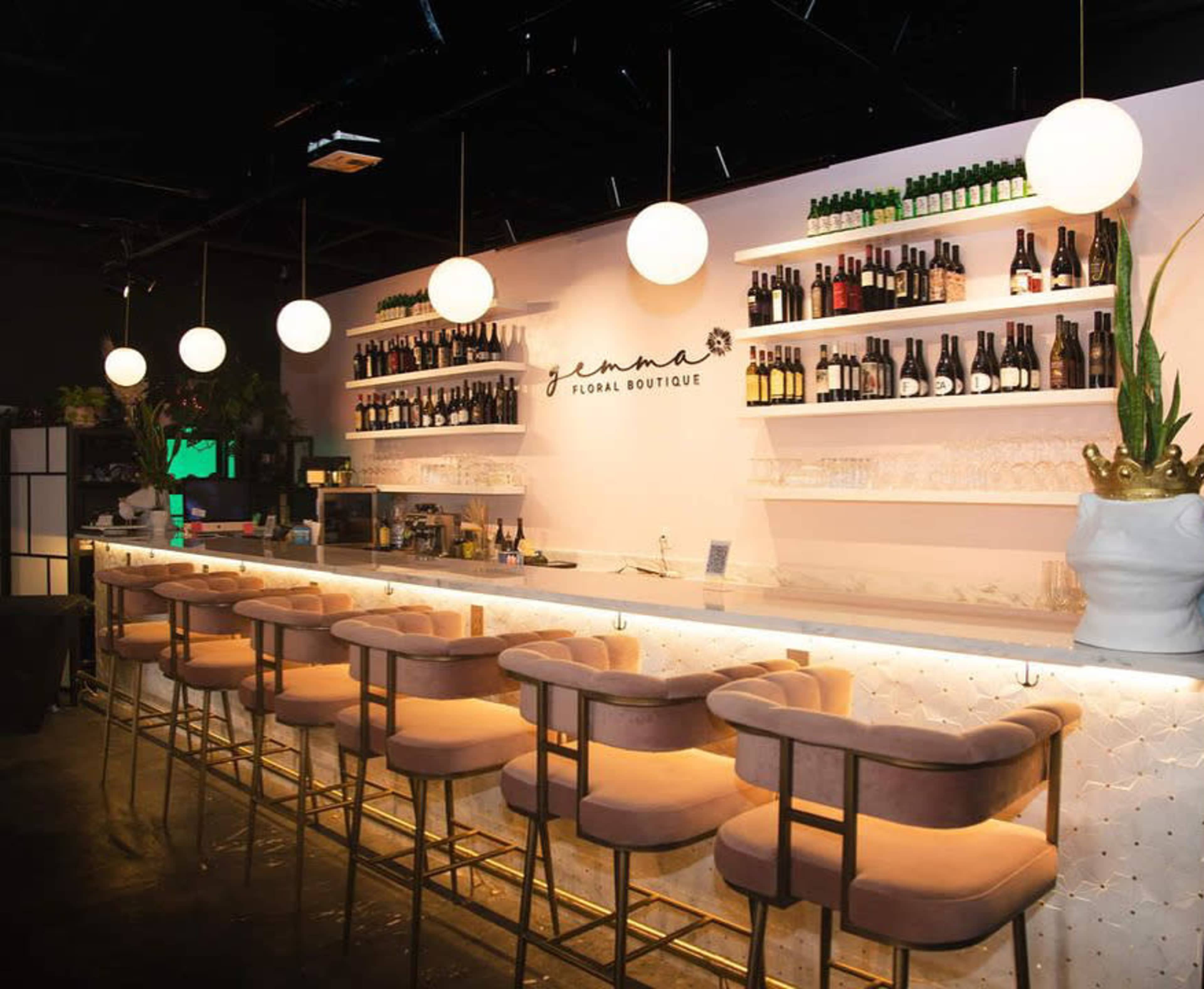 The image shows a modern bar area featuring a white wall with shelves displaying various bottles, illuminated by round pendant lights, and adorned with plush pink stools along the counter.