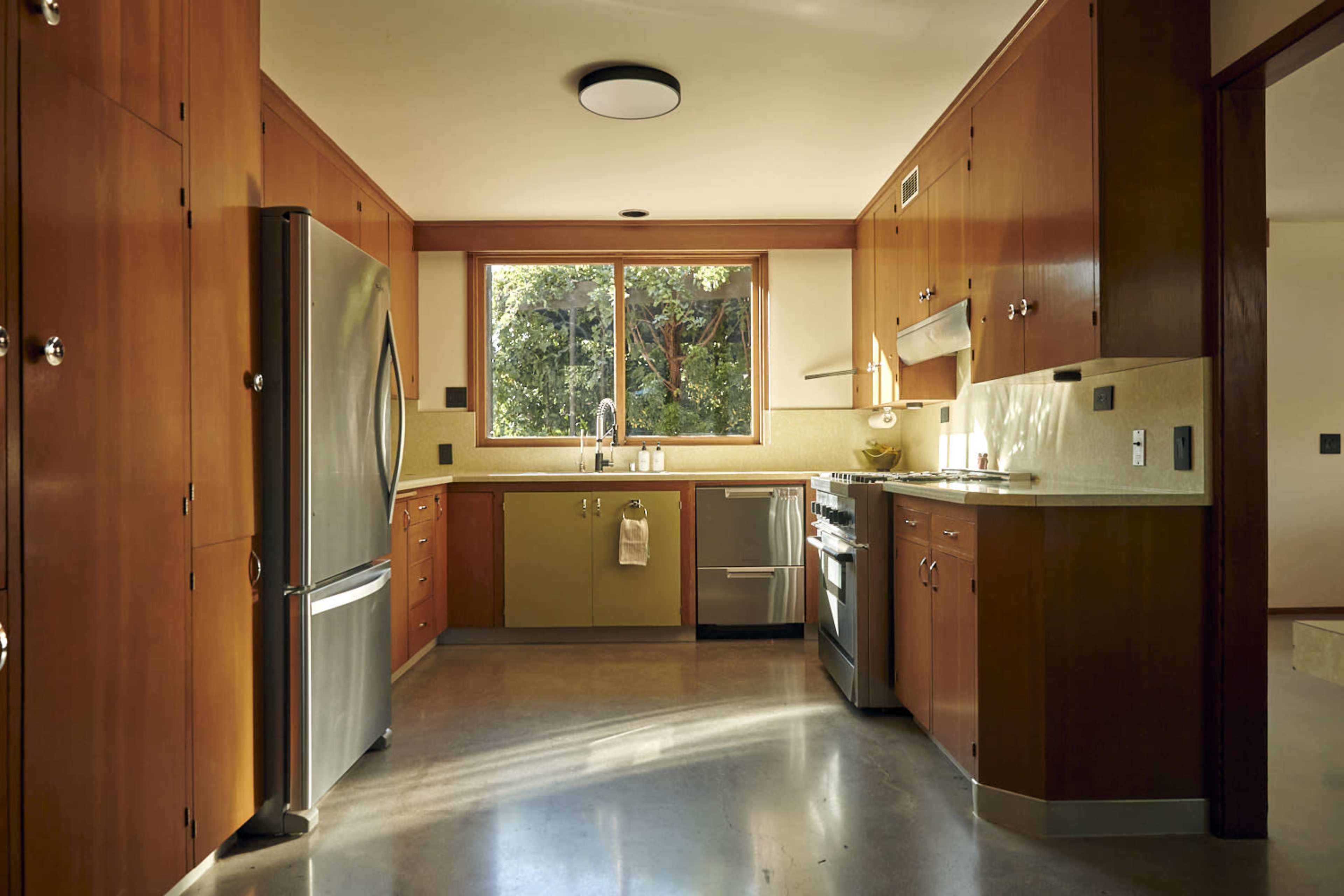 The image shows a modern kitchen with wooden cabinetry, stainless steel appliances, and a large window allowing natural light to enter.