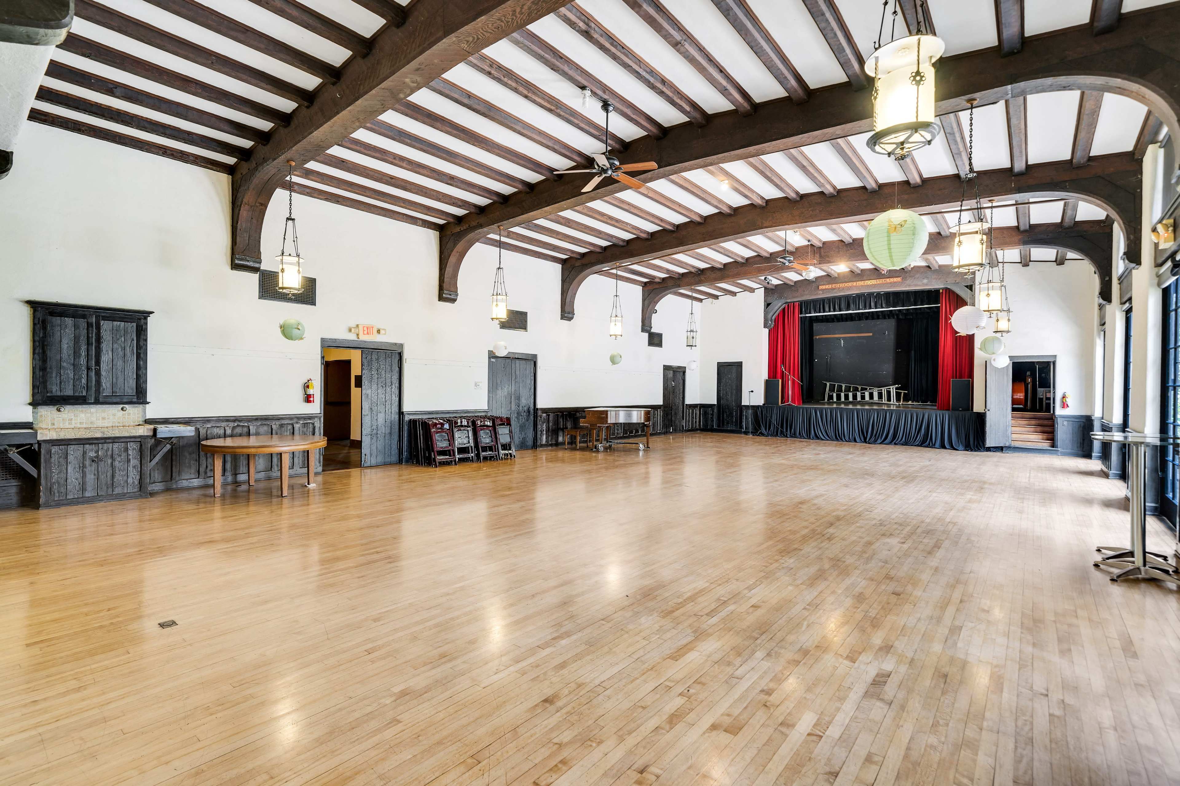 The image shows a spacious indoor hall featuring wooden beams, a polished floor, and a raised stage with red curtains at one end.