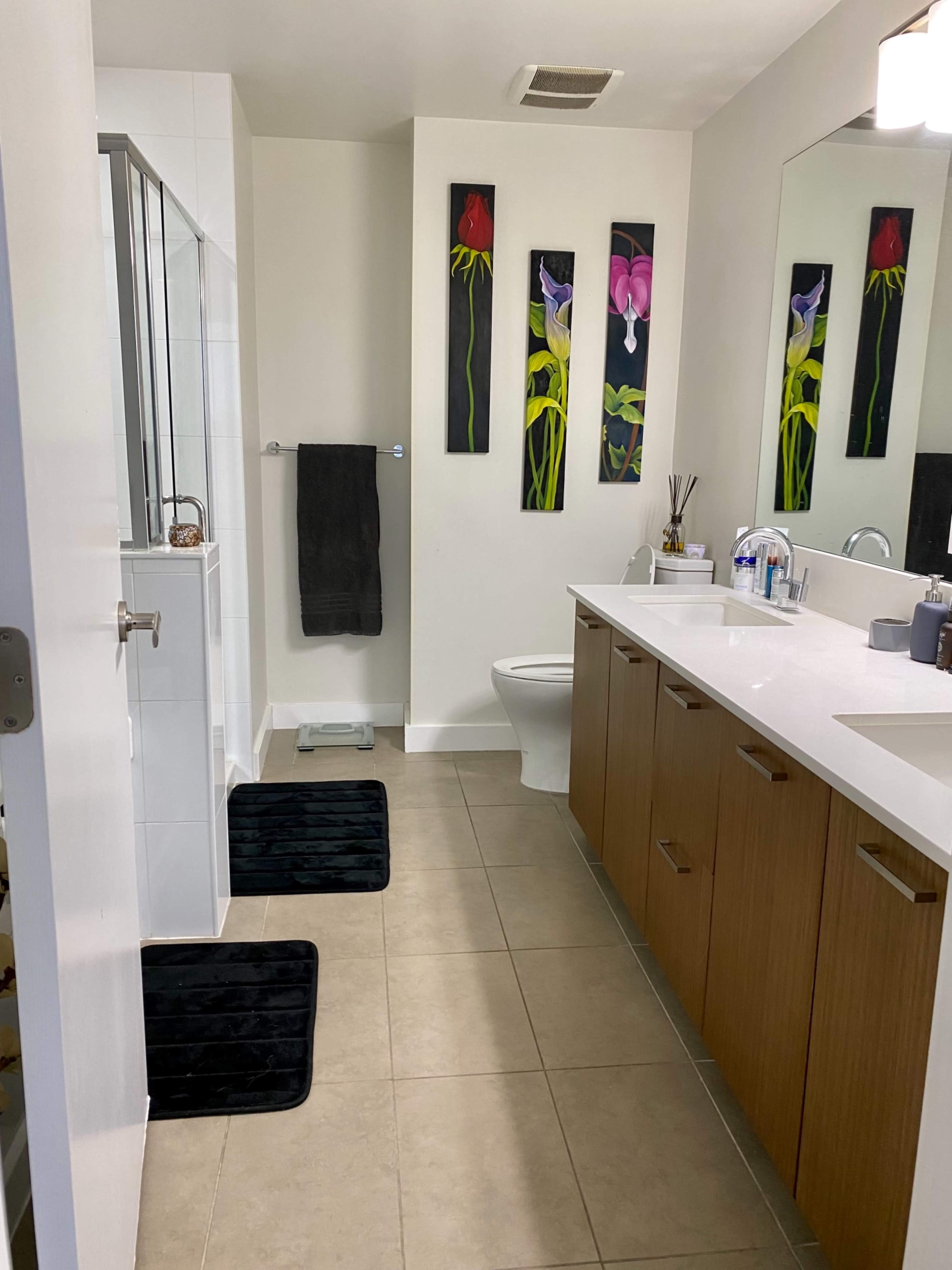 A modern bathroom features a walk-in shower, a toilet, a double vanity with wooden cabinets, and floral wall art.