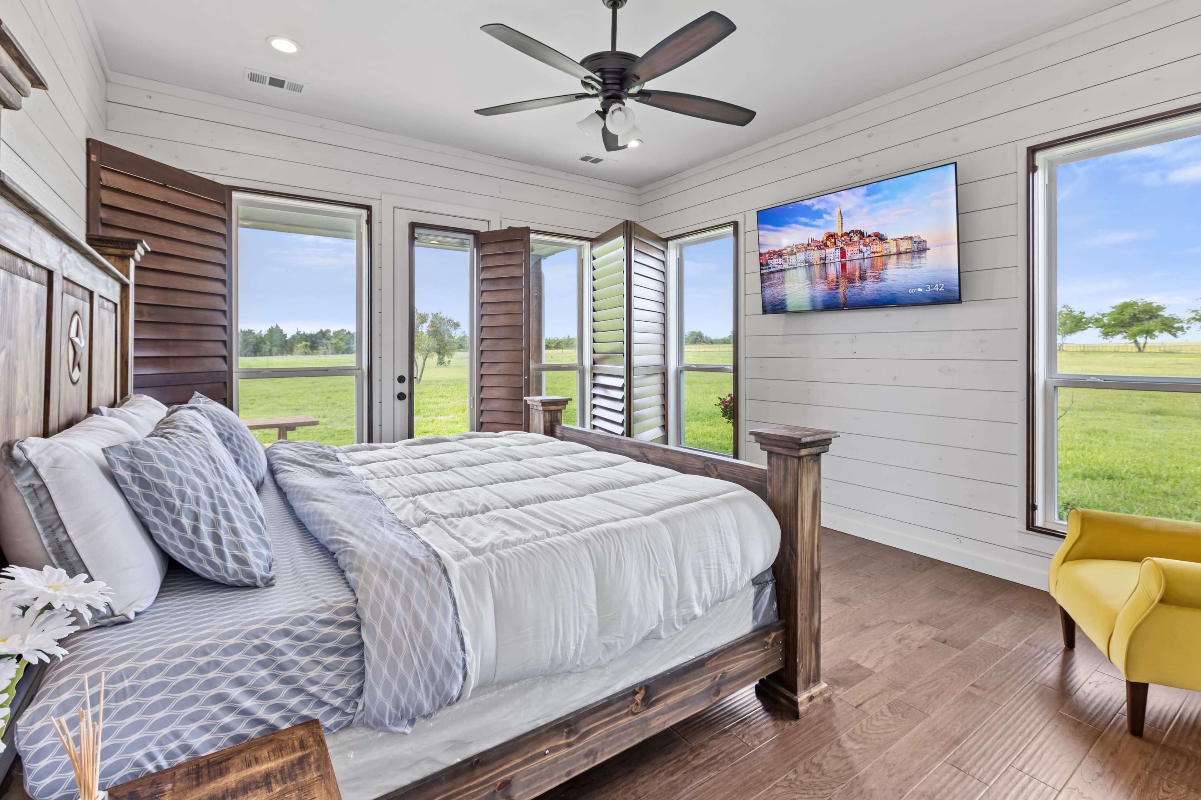 A clean and bright bedroom features a wooden bed, large windows with open shutters, and a wall-mounted television overlooking a grassy landscape.