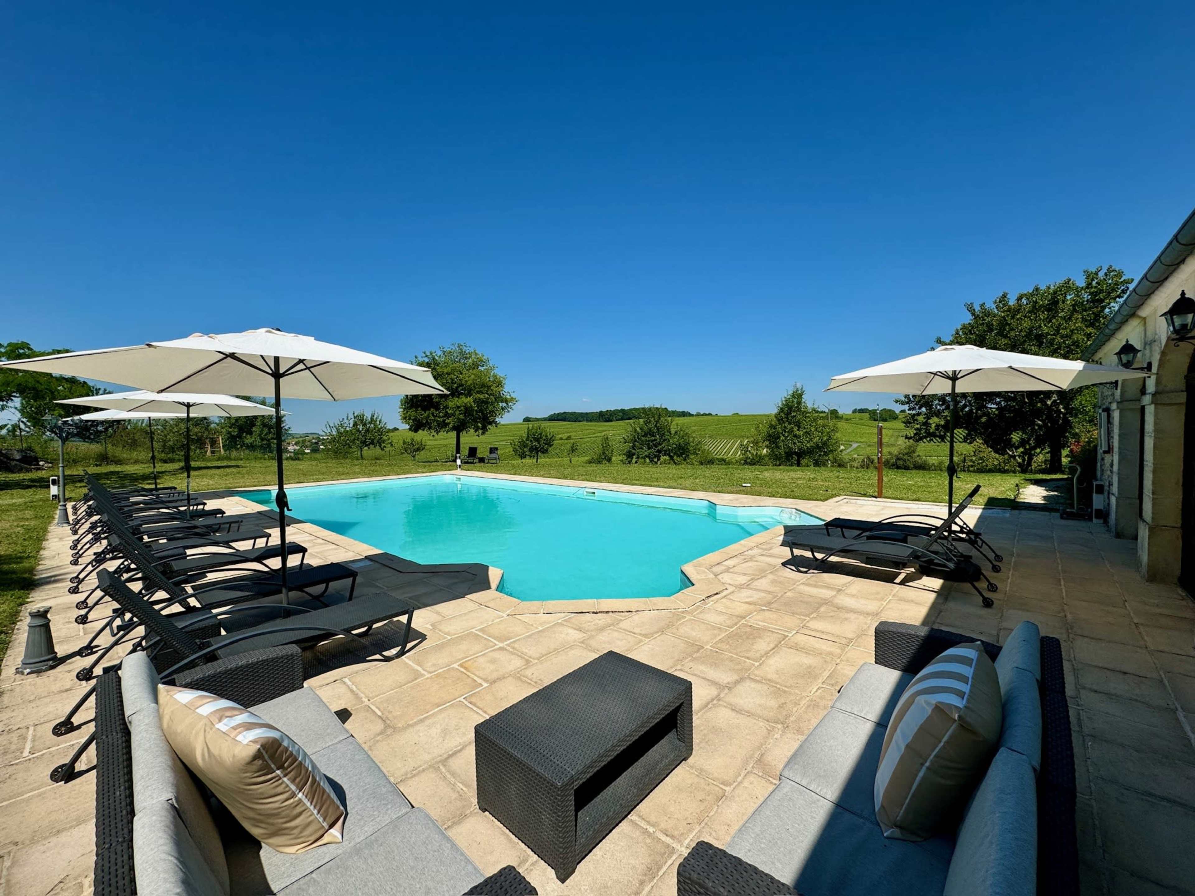 The image shows a swimming pool surrounded by lounge chairs and umbrellas, with green fields and trees in the background under a clear blue sky.