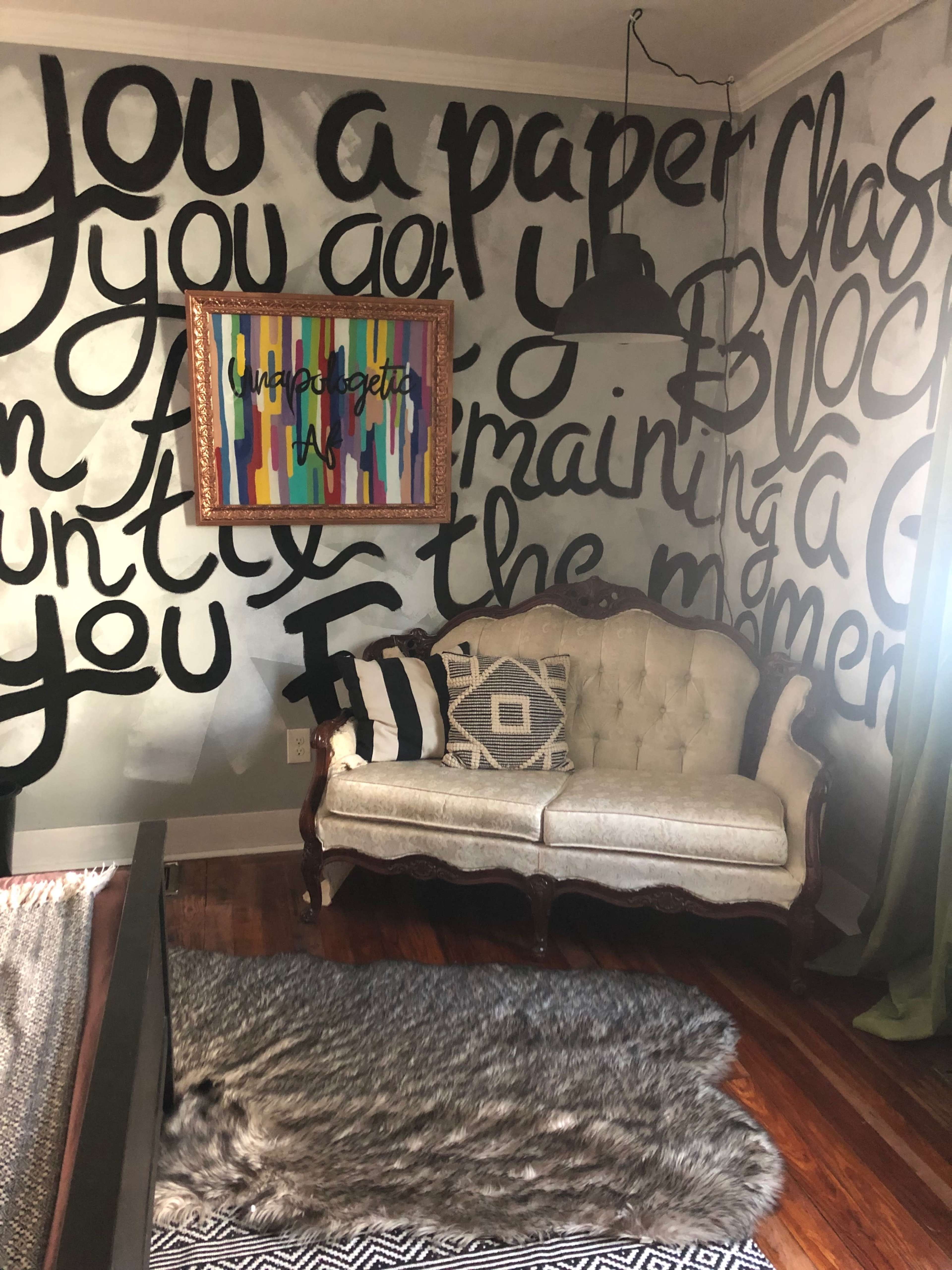 A cozy room with a vintage couch, a large text mural on the wall, a colorful framed artwork, and a soft rug on wooden flooring.