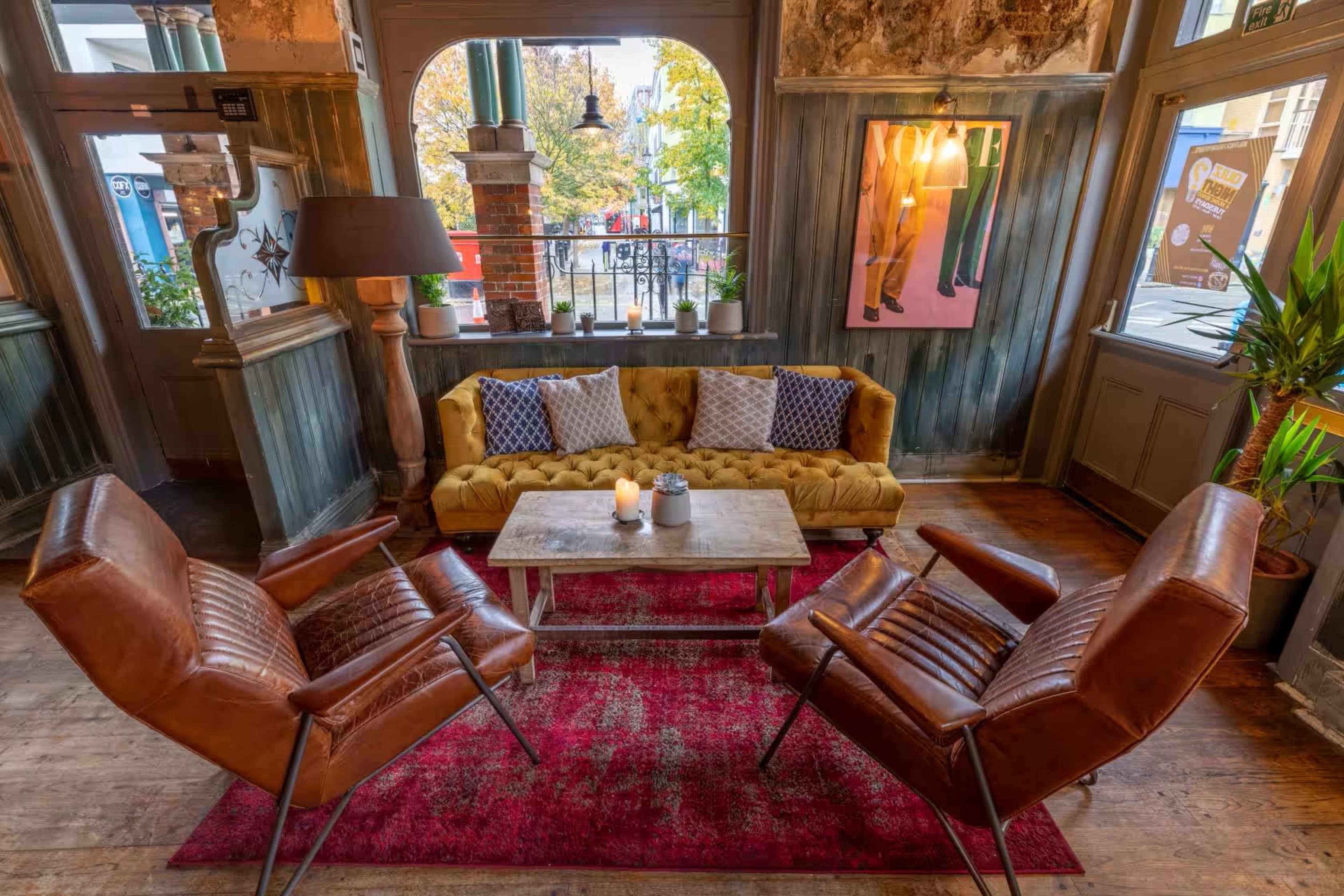 The scene features a cozy lounge area with a yellow sofa and two brown chairs positioned around a small table on a red rug, illuminated by natural light from large windows.