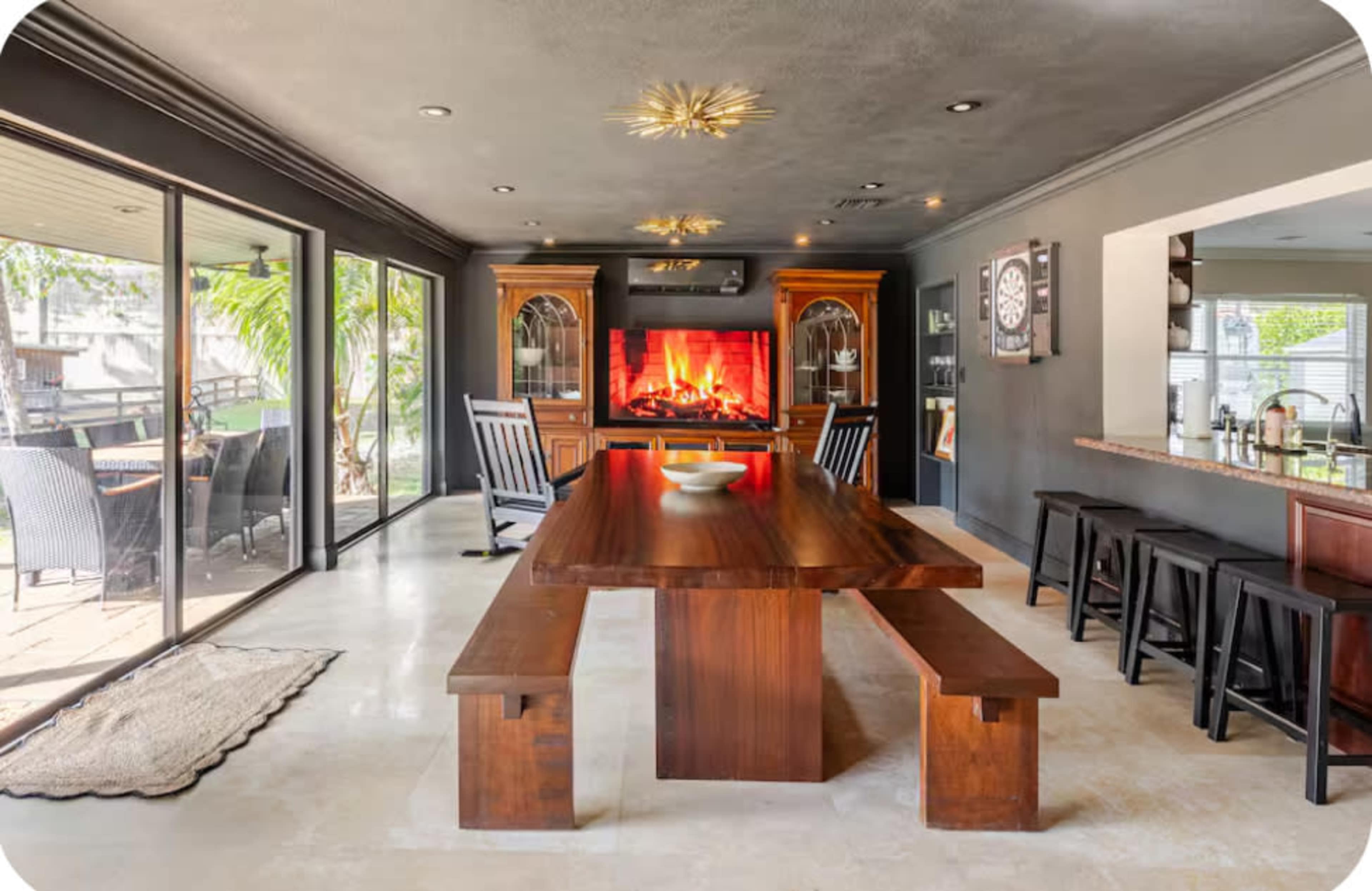 The image shows a modern dining area with a large wooden table surrounded by benches, facing a wall-mounted fireplace and large windows that open to an outdoor space.