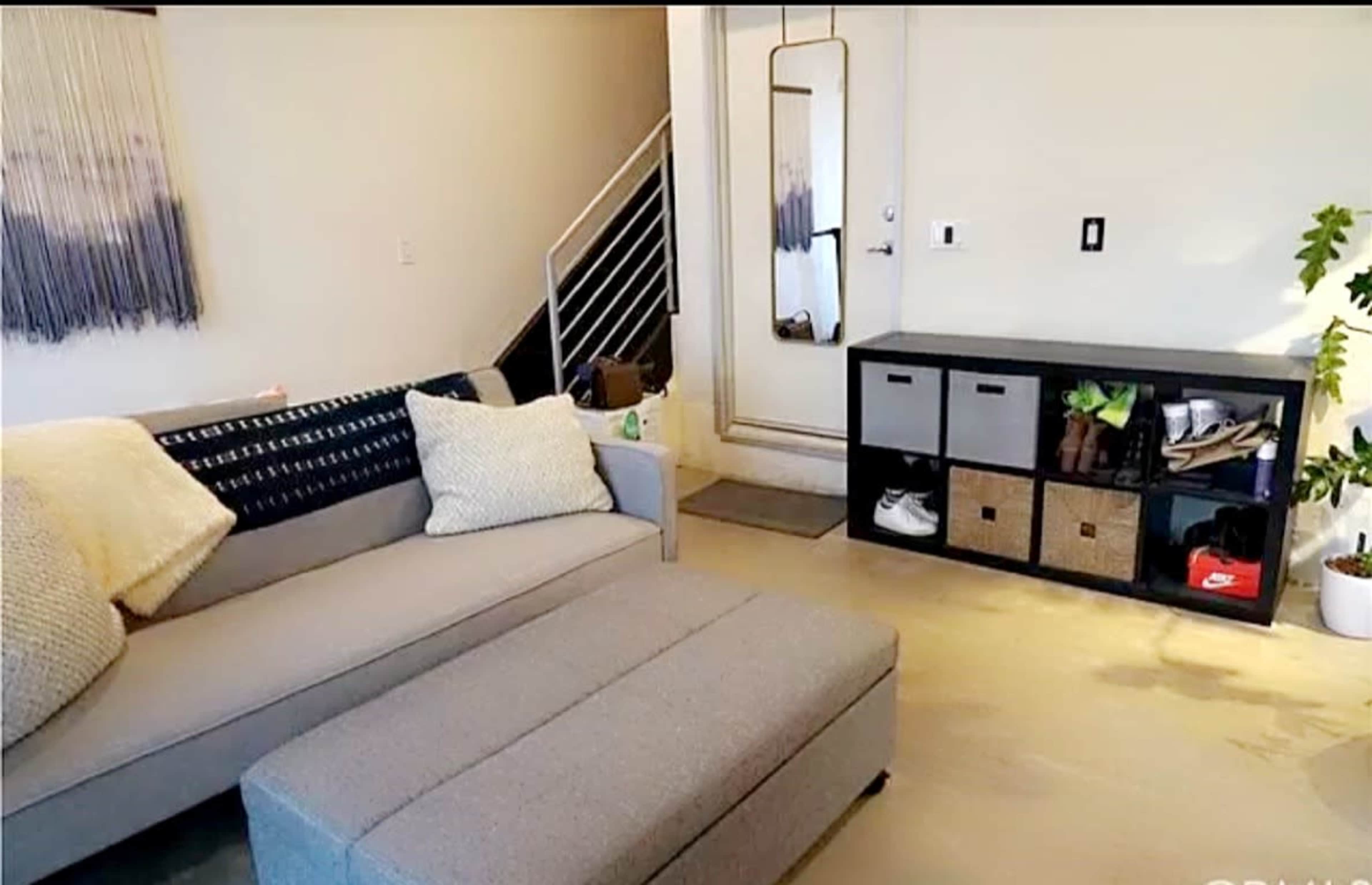 A modern living room featuring a gray couch with decorative pillows, an ottoman, and a storage unit with baskets and shoes against a wall.