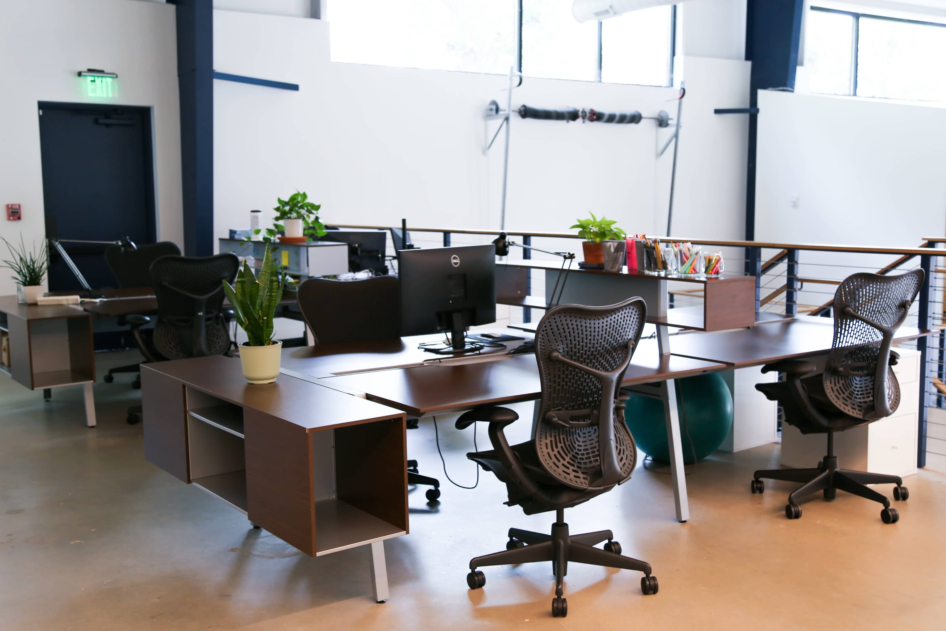 The image shows a modern office space featuring several desks with ergonomic chairs, plants, and computer equipment arranged in an open layout.
