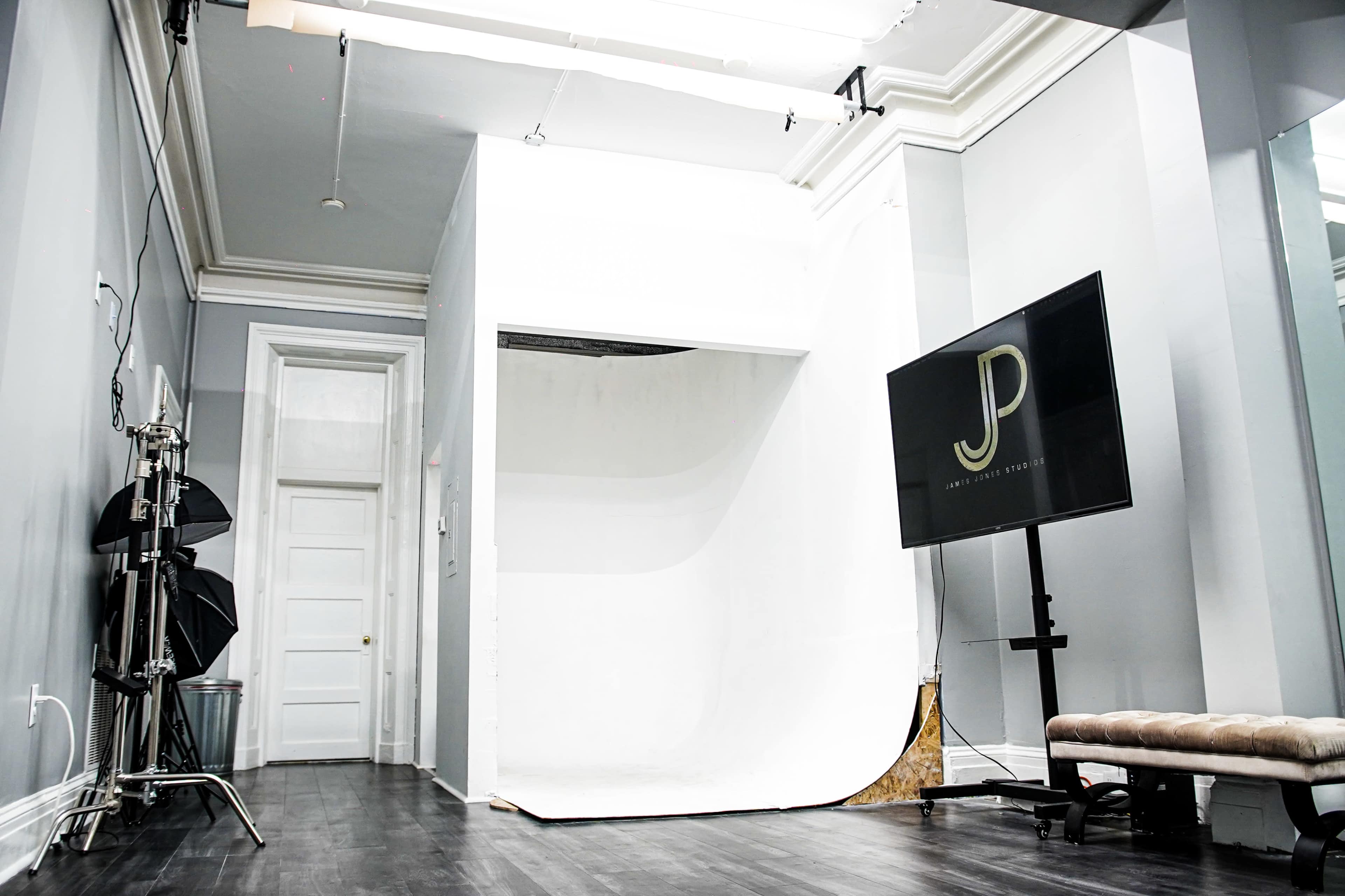 A minimalist photography studio with a white backdrop, a large flat-screen television displaying the initials "JP," and studio lighting equipment positioned nearby.