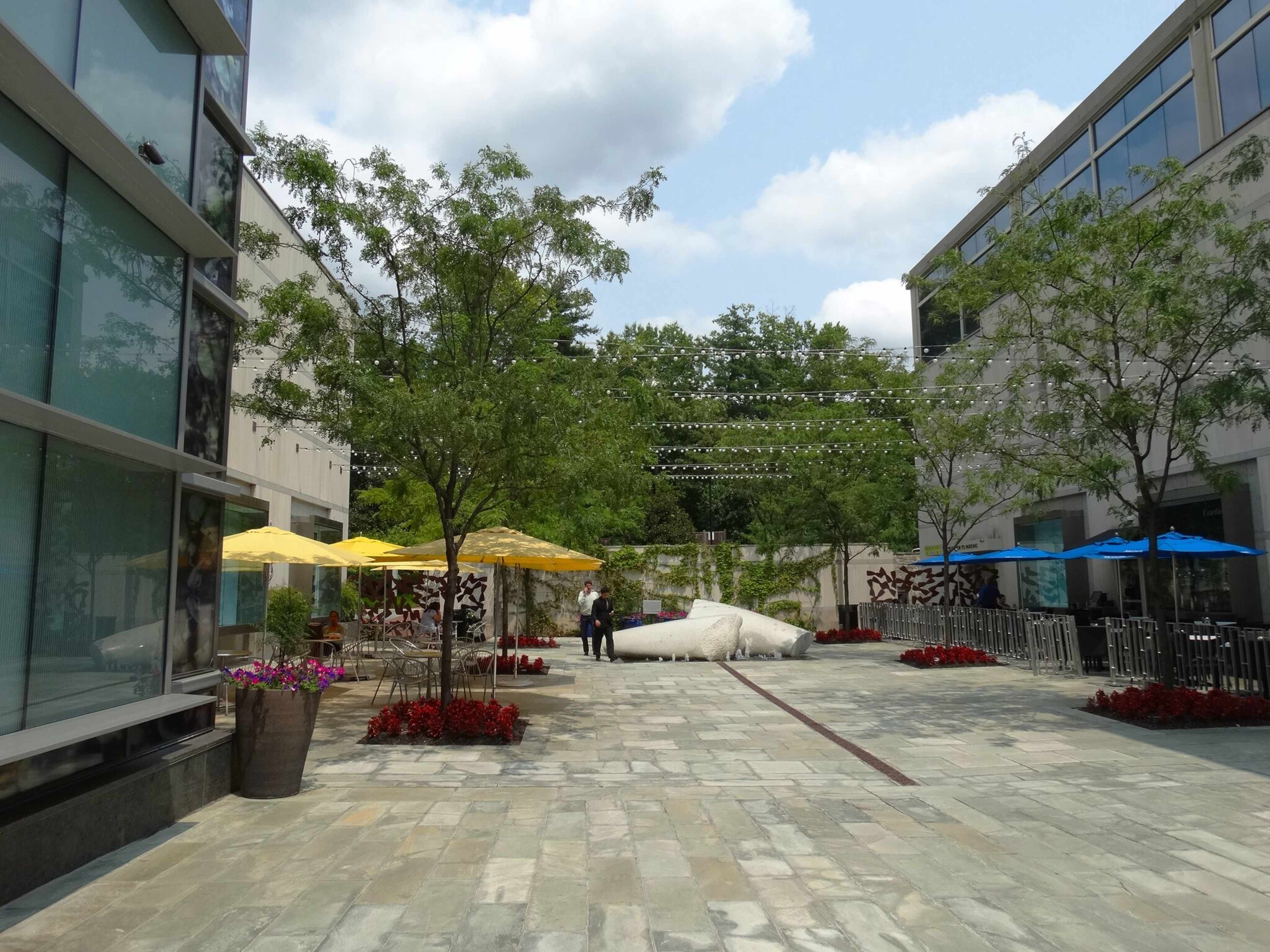 A landscaped outdoor courtyard with stone paving, decorative trees, colorful umbrellas, and seating areas between two modern buildings.