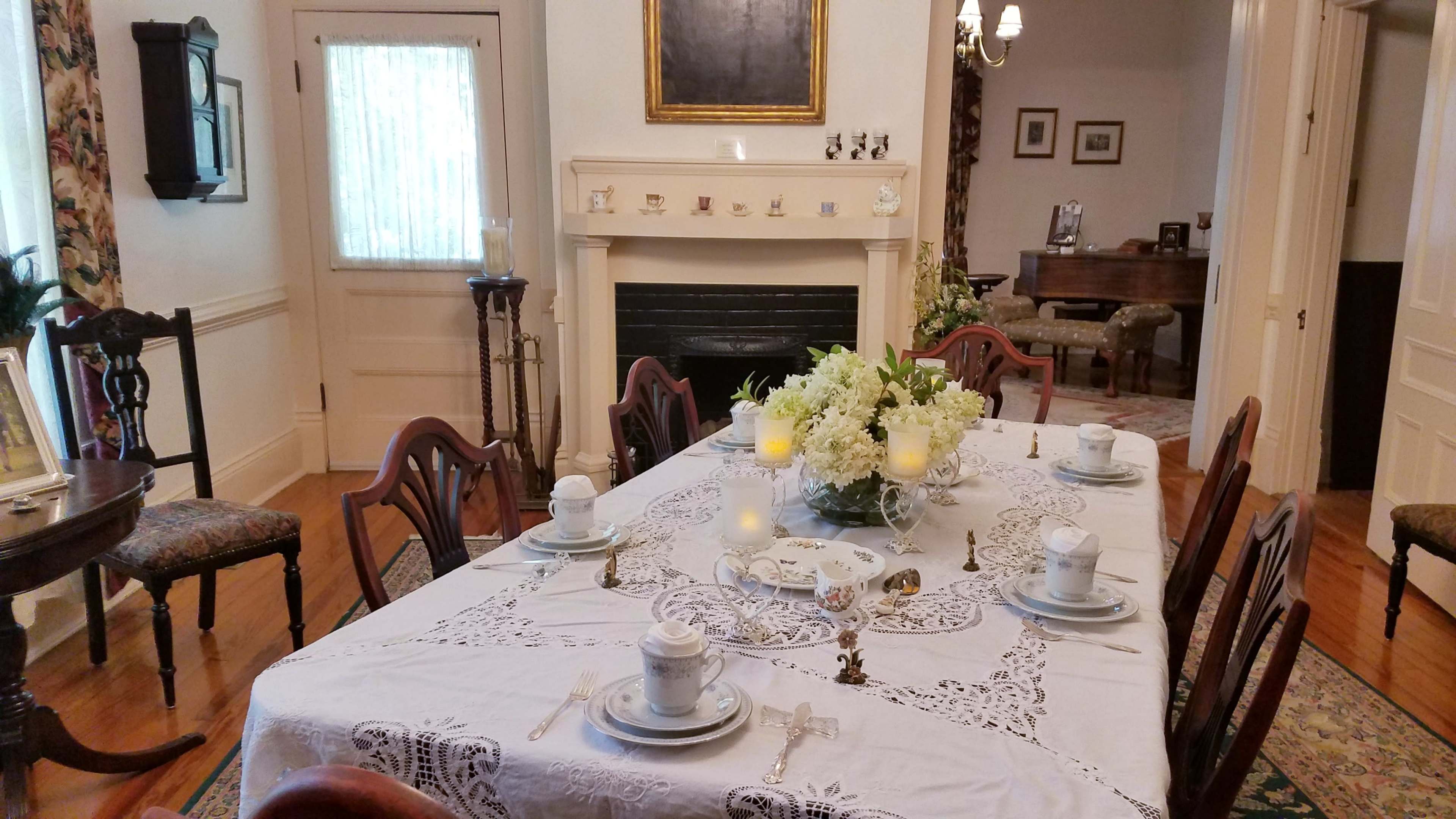 A dining room is set with a white lace tablecloth and fine china, featuring a floral centerpiece and decorative candles.