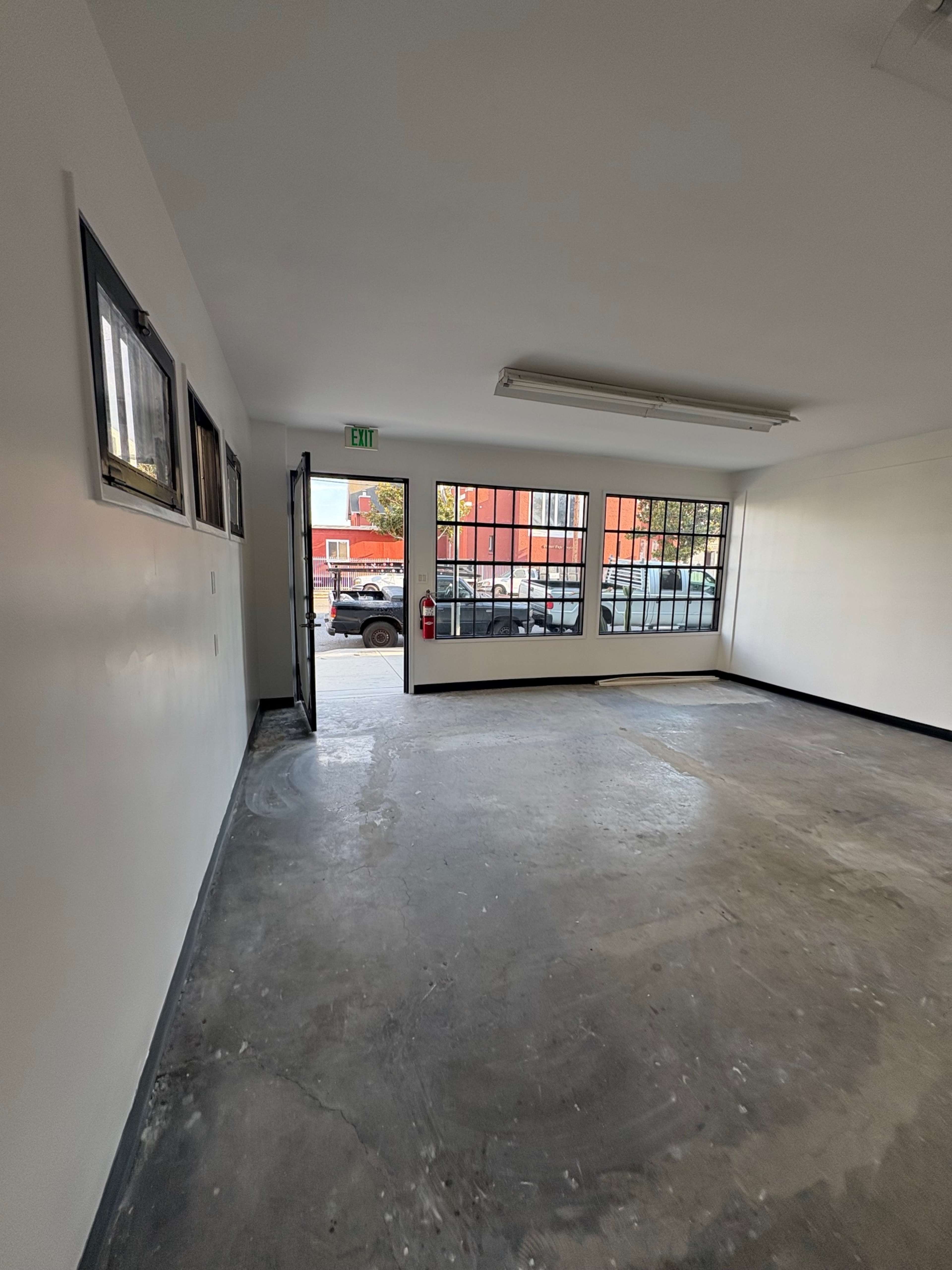 The image shows an empty room with bare concrete floors and large windows, providing a view of the outside.
