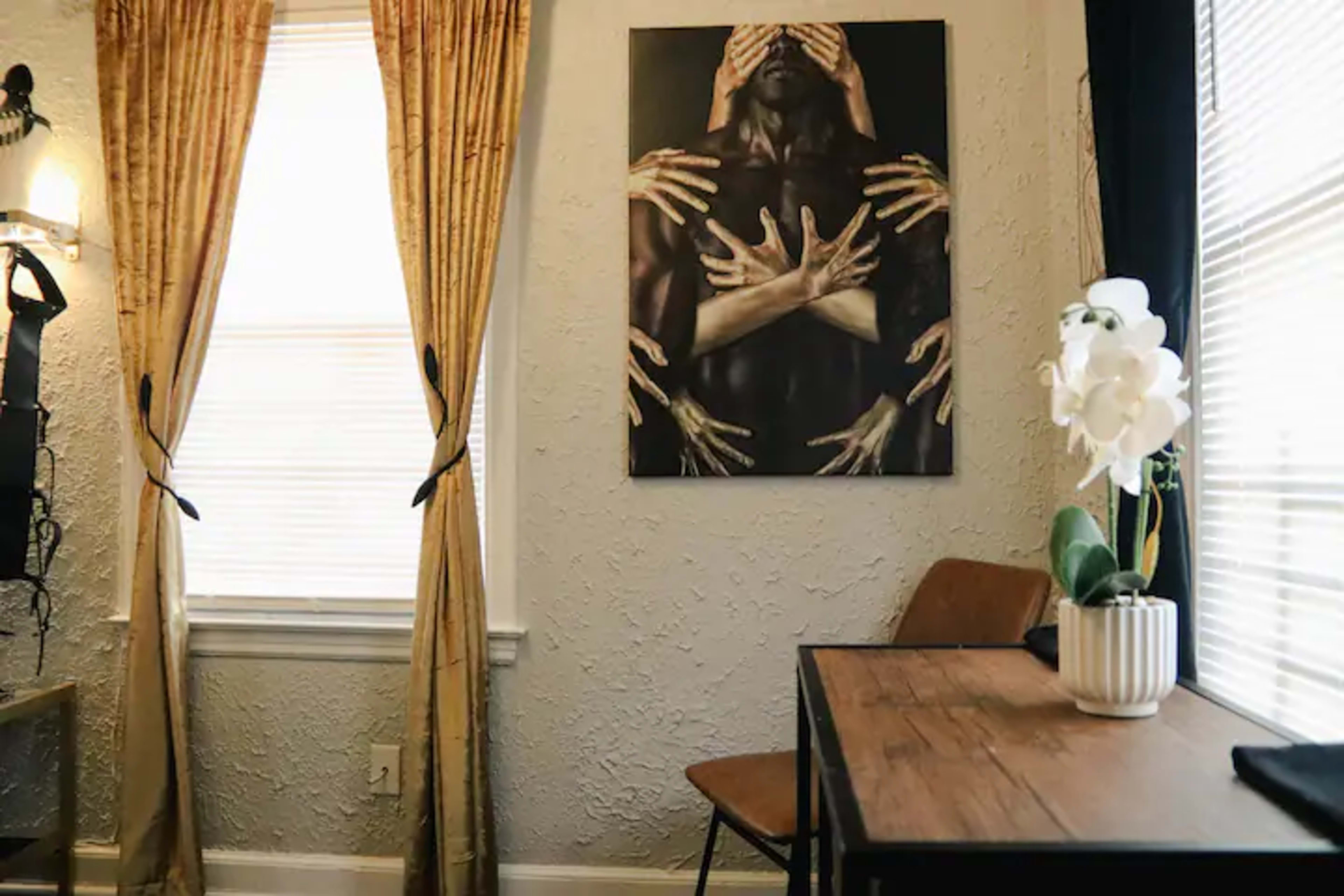 A small dining area features a wooden table and a chair beside a wall adorned with a large artwork depicting multiple hands covering a figure's face, with curtains framing a window.