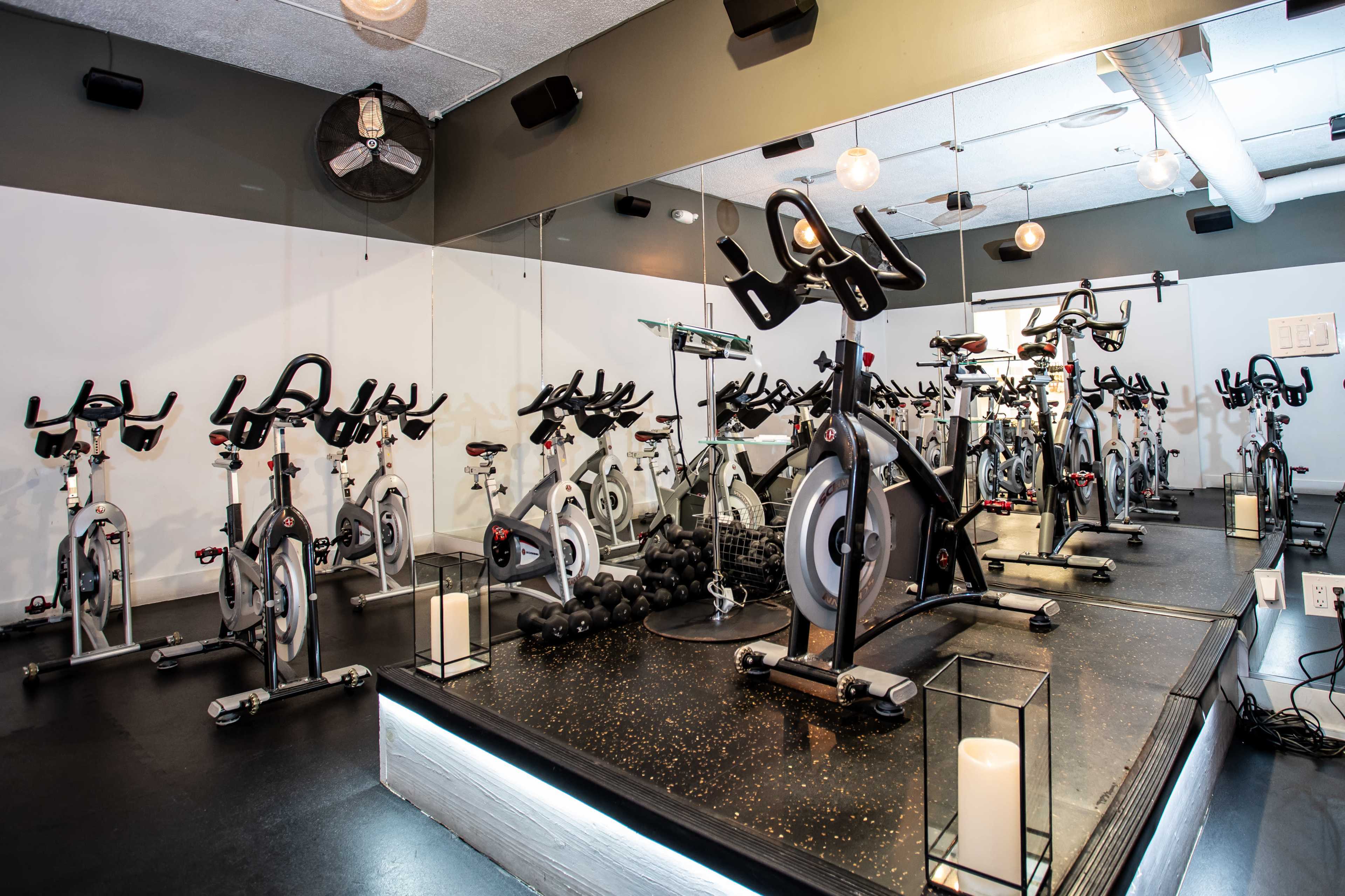 The image shows a fitness studio with multiple stationary bicycles arranged in rows, facing a mirrored wall, and equipped with some weights and ambient lighting.