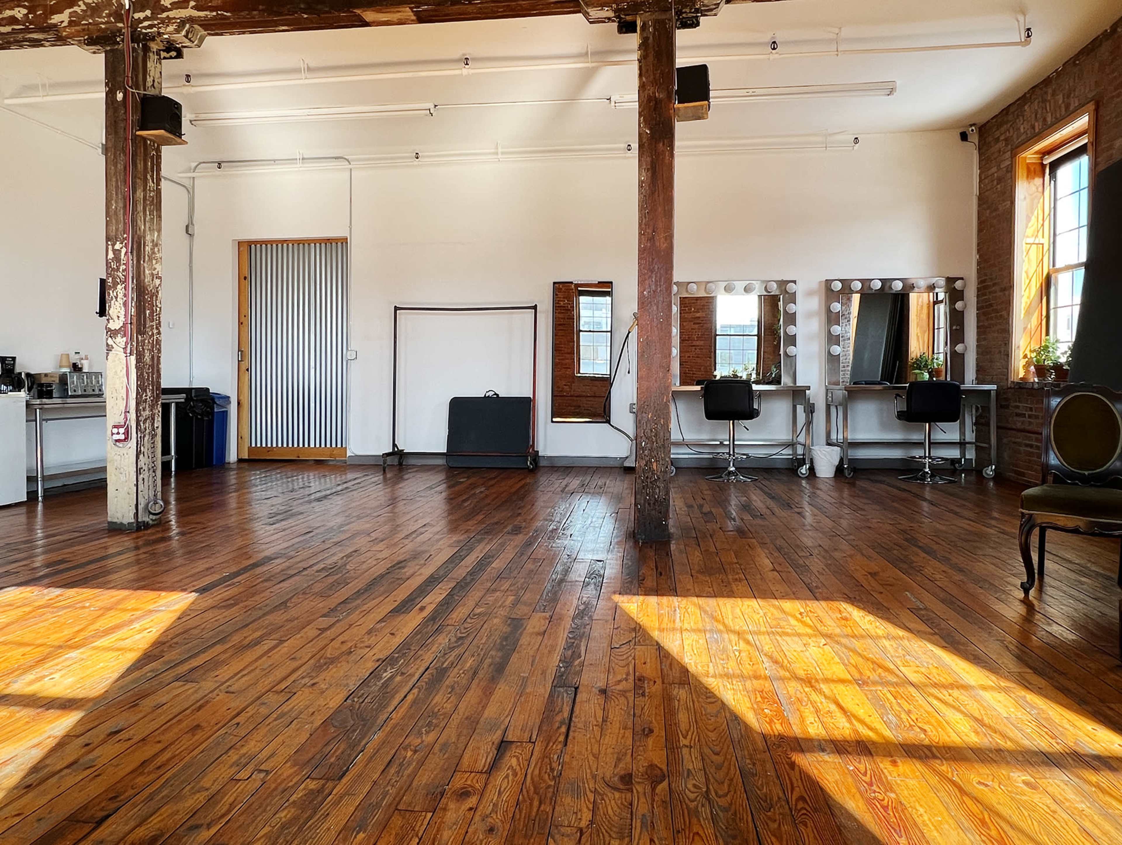 An empty studio space features wooden flooring, large windows, and makeup mirrors along one wall.