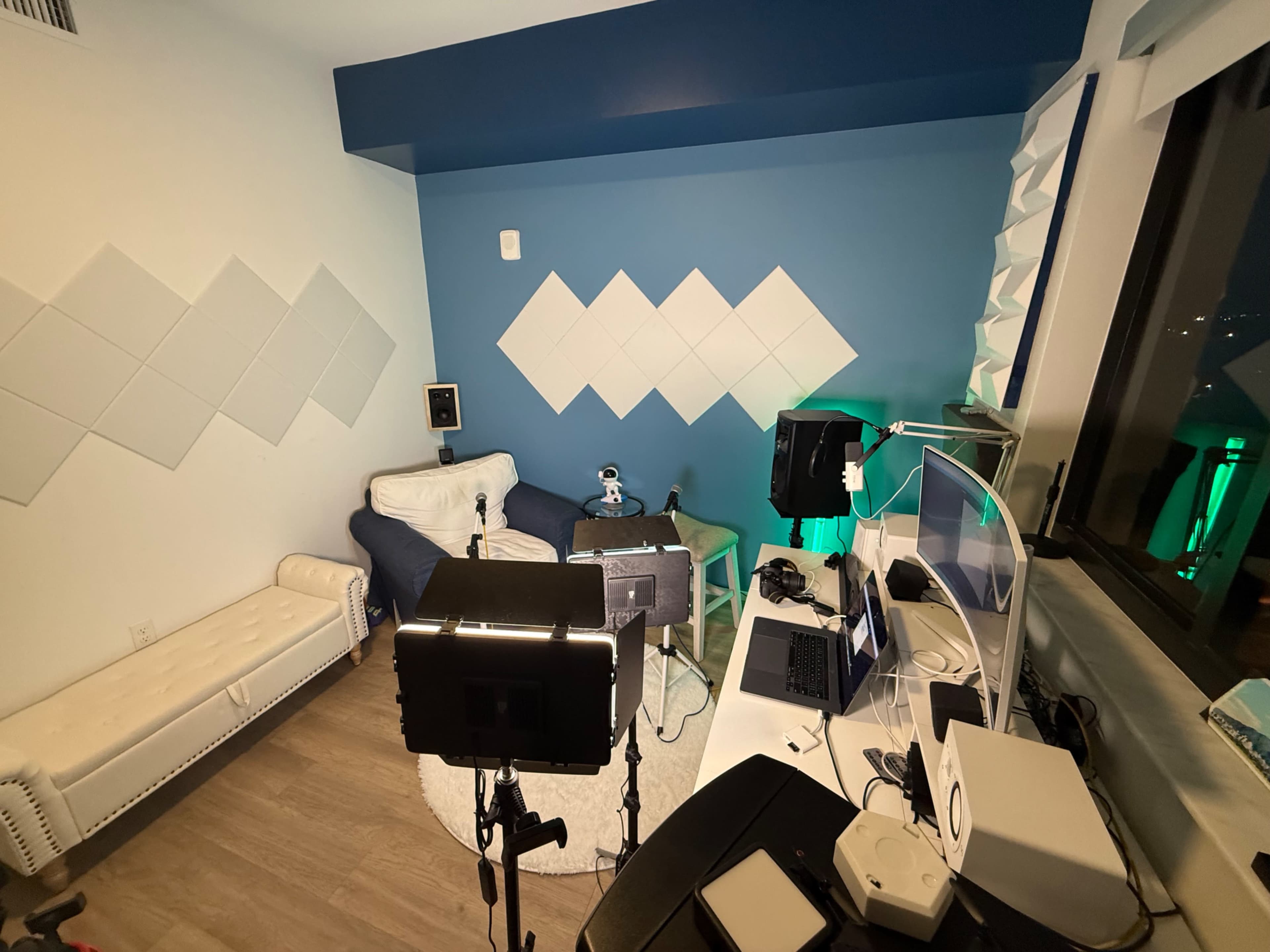 The image shows a well-lit recording studio with a white couch, green and white walls, and various equipment including microphones, cameras, and a laptop set up on a desk.