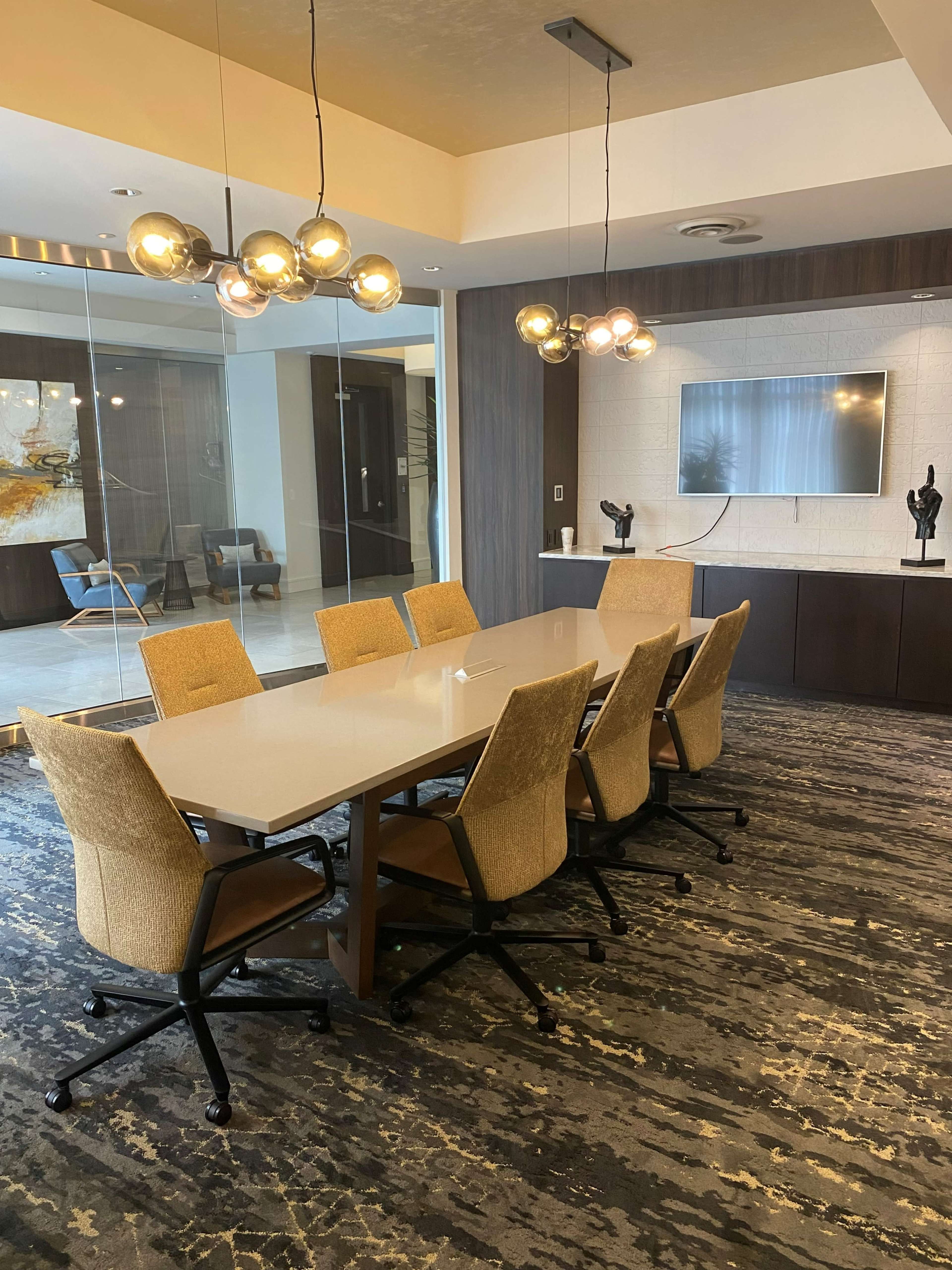 A modern conference room features a long table surrounded by six beige chairs, with a television mounted on the wall and decorative sculptures on a side cabinet.