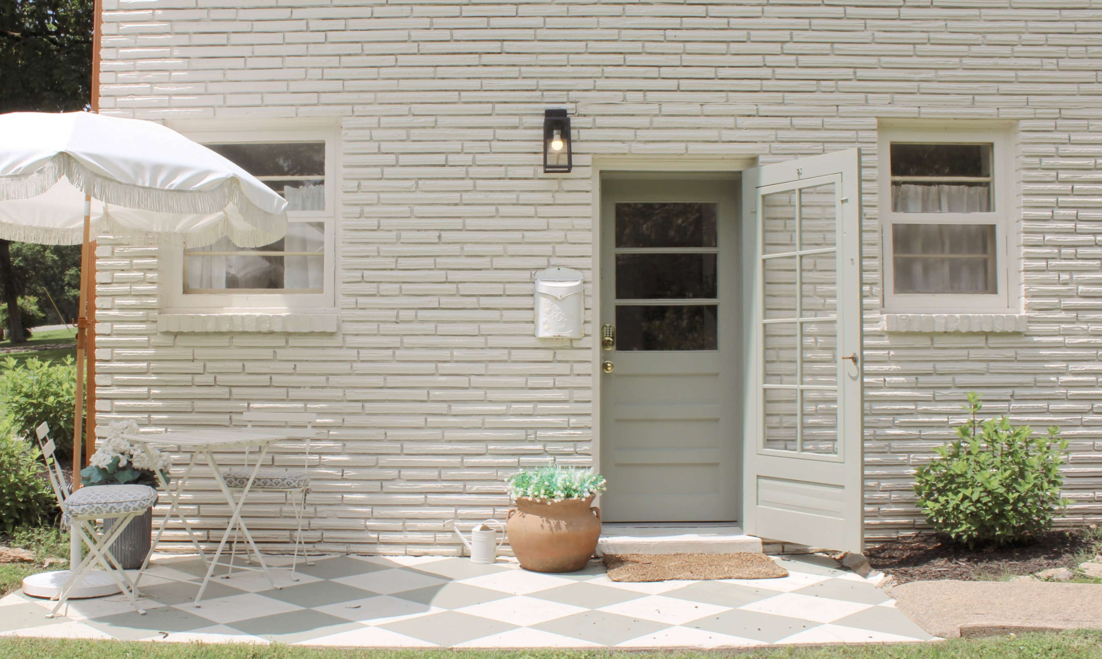 A light-colored brick building features an open door with a small table and chairs beside a large potted plant on a patterned porch.