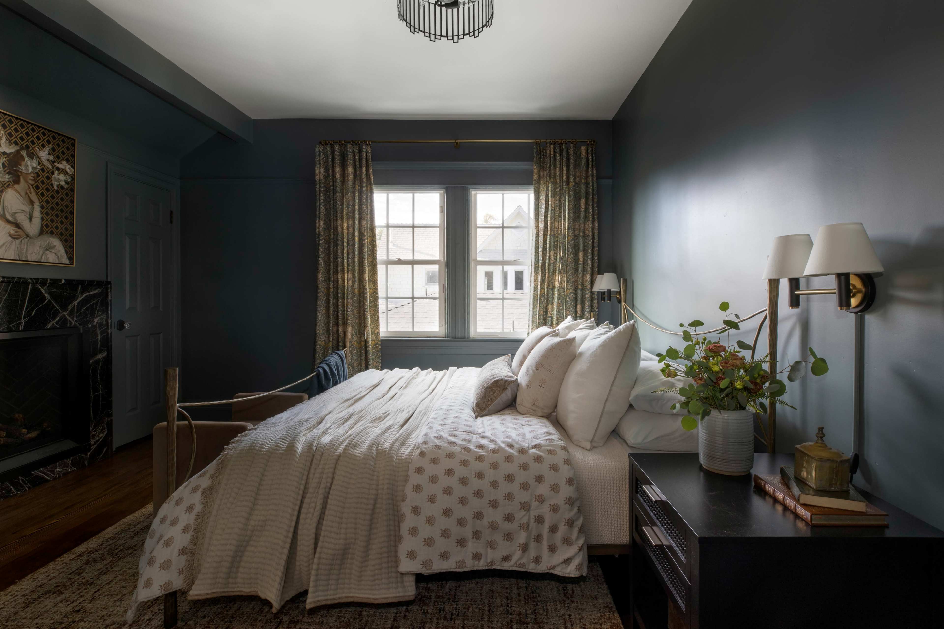 A neatly arranged bedroom with a large window, a bed with white linens, and a small seating area near a fireplace.