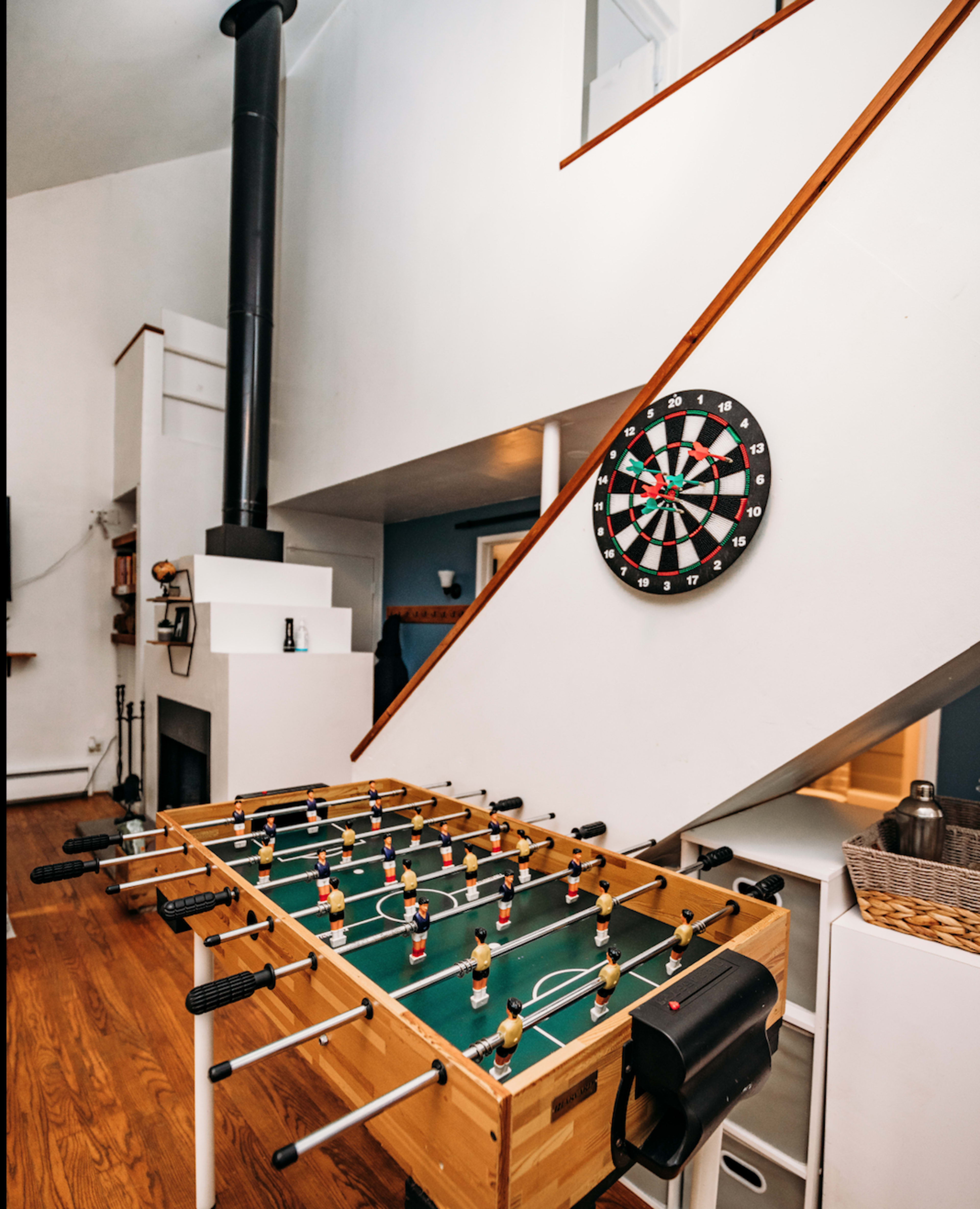 A foosball table is positioned in a room with a staircase and a dartboard mounted on the wall.