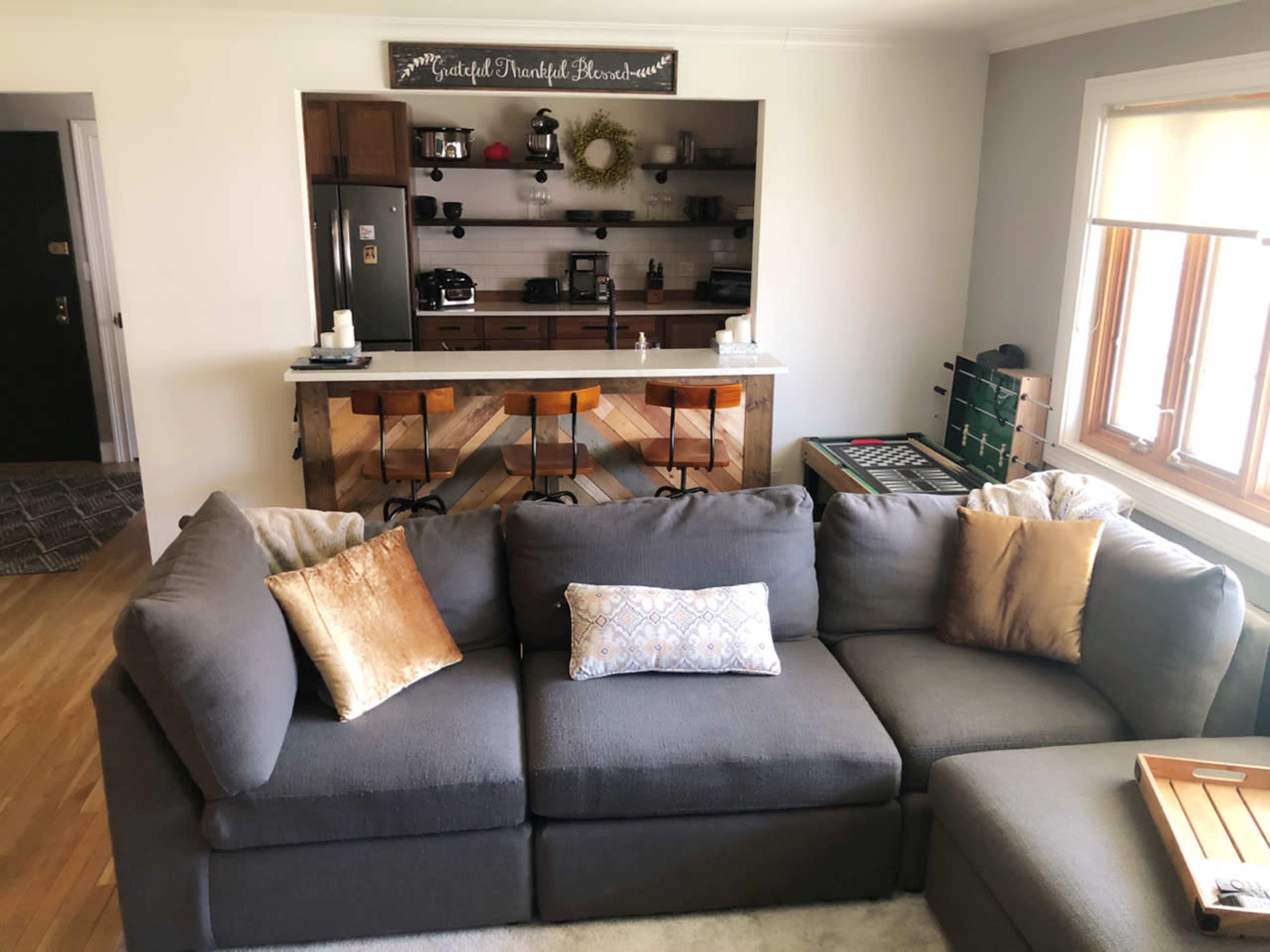 The image shows a cozy living room with a dark gray sectional sofa, a small wooden coffee table, and a view of a kitchen area with a bar and various appliances in the background.