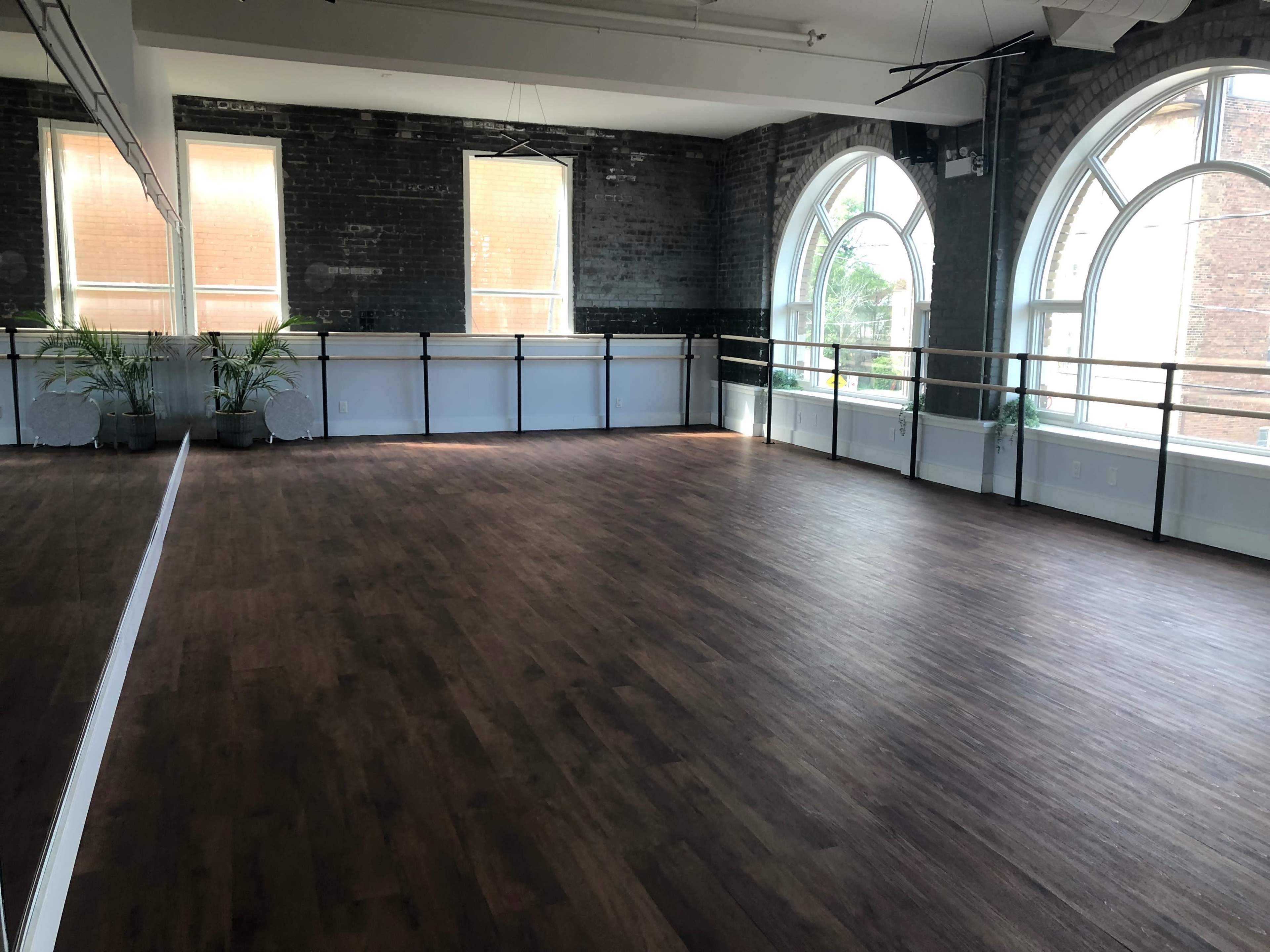The image shows a spacious dance studio with large windows, exposed brick walls, and a wooden floor.