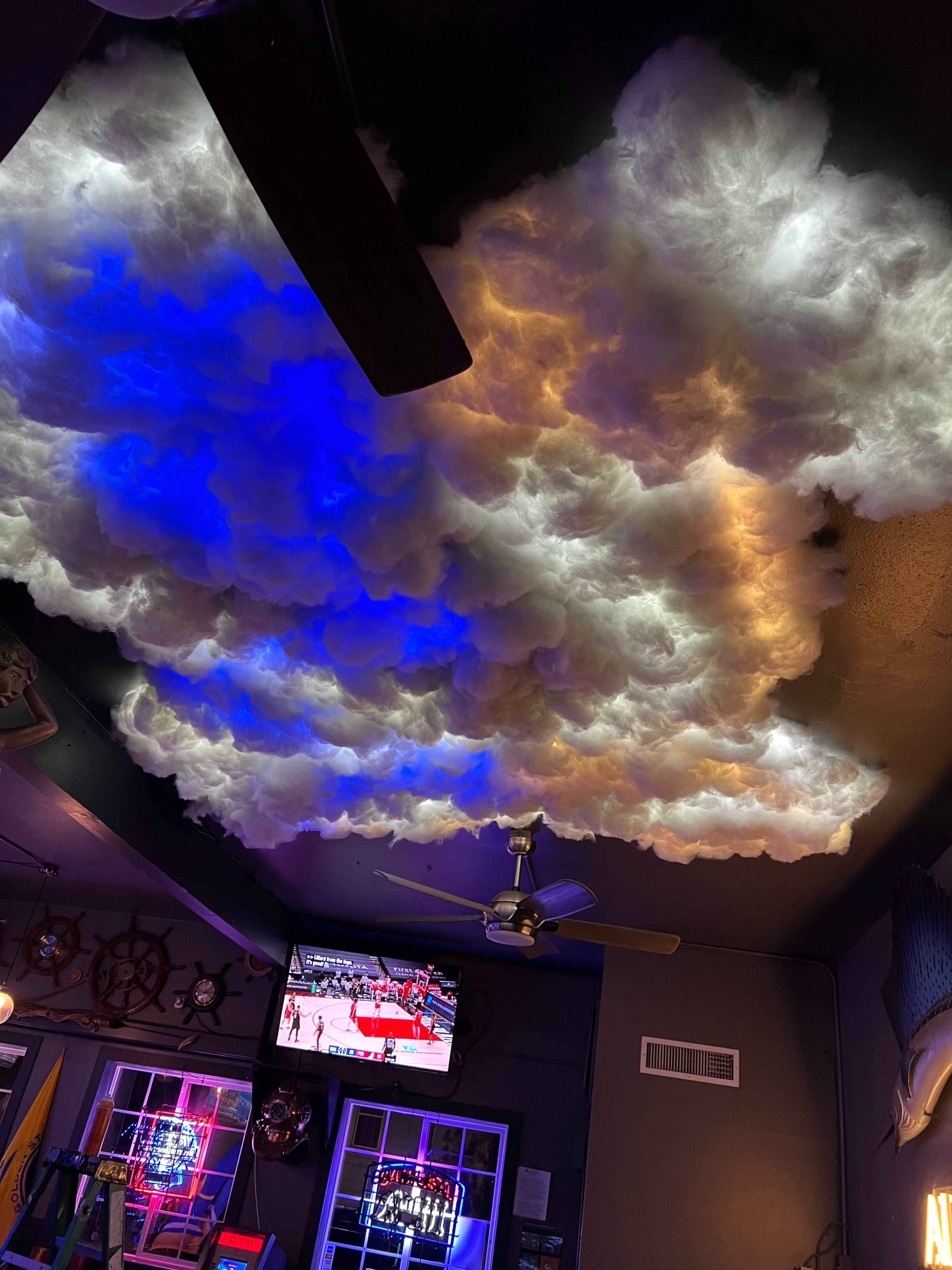 The ceiling features artificial clouds with blue and orange lighting, while a television displays a basketball game in the background.