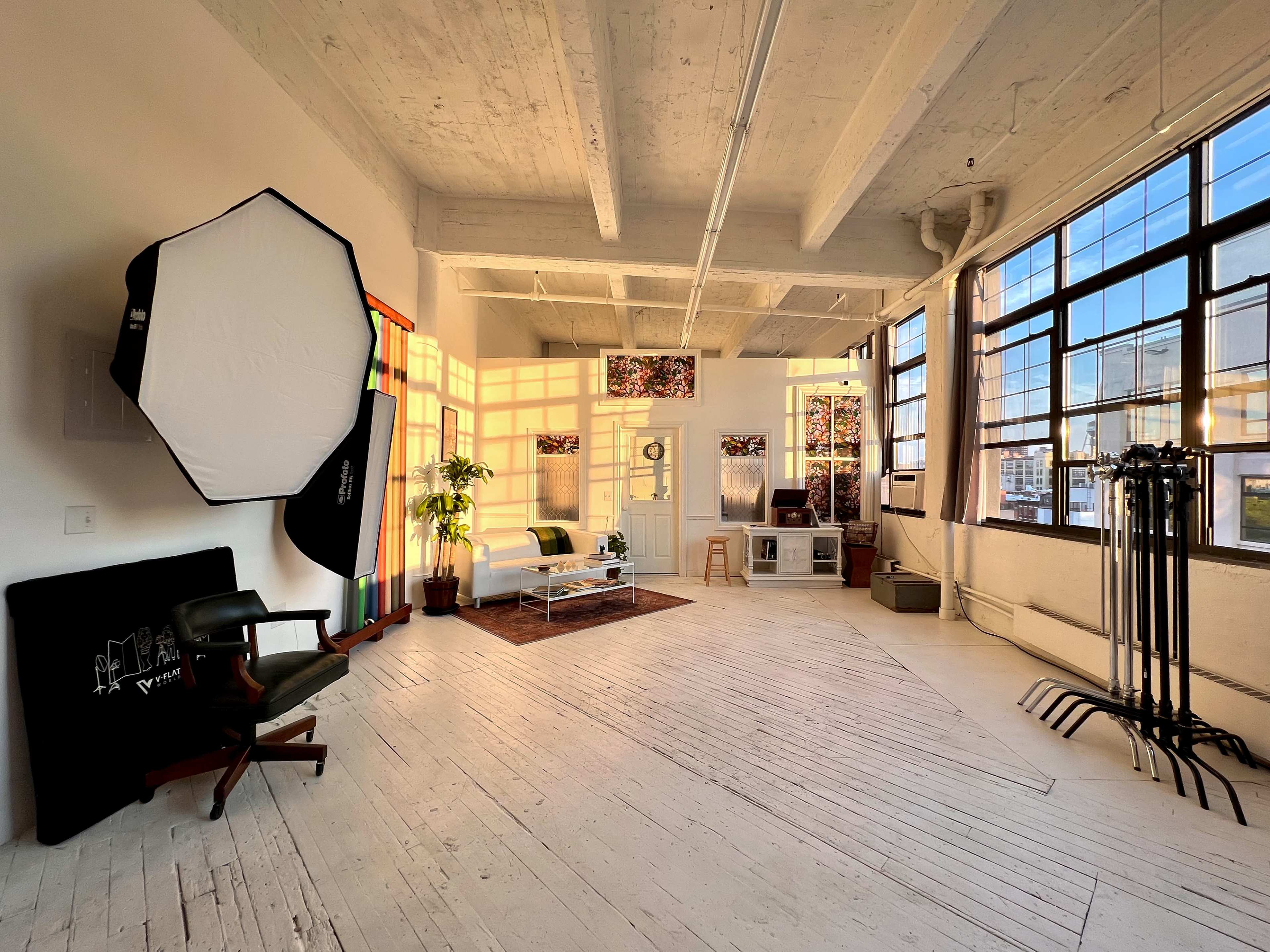 A spacious brightly lit studio with large windows, featuring a white couch, a rug, and various pieces of equipment along the walls.