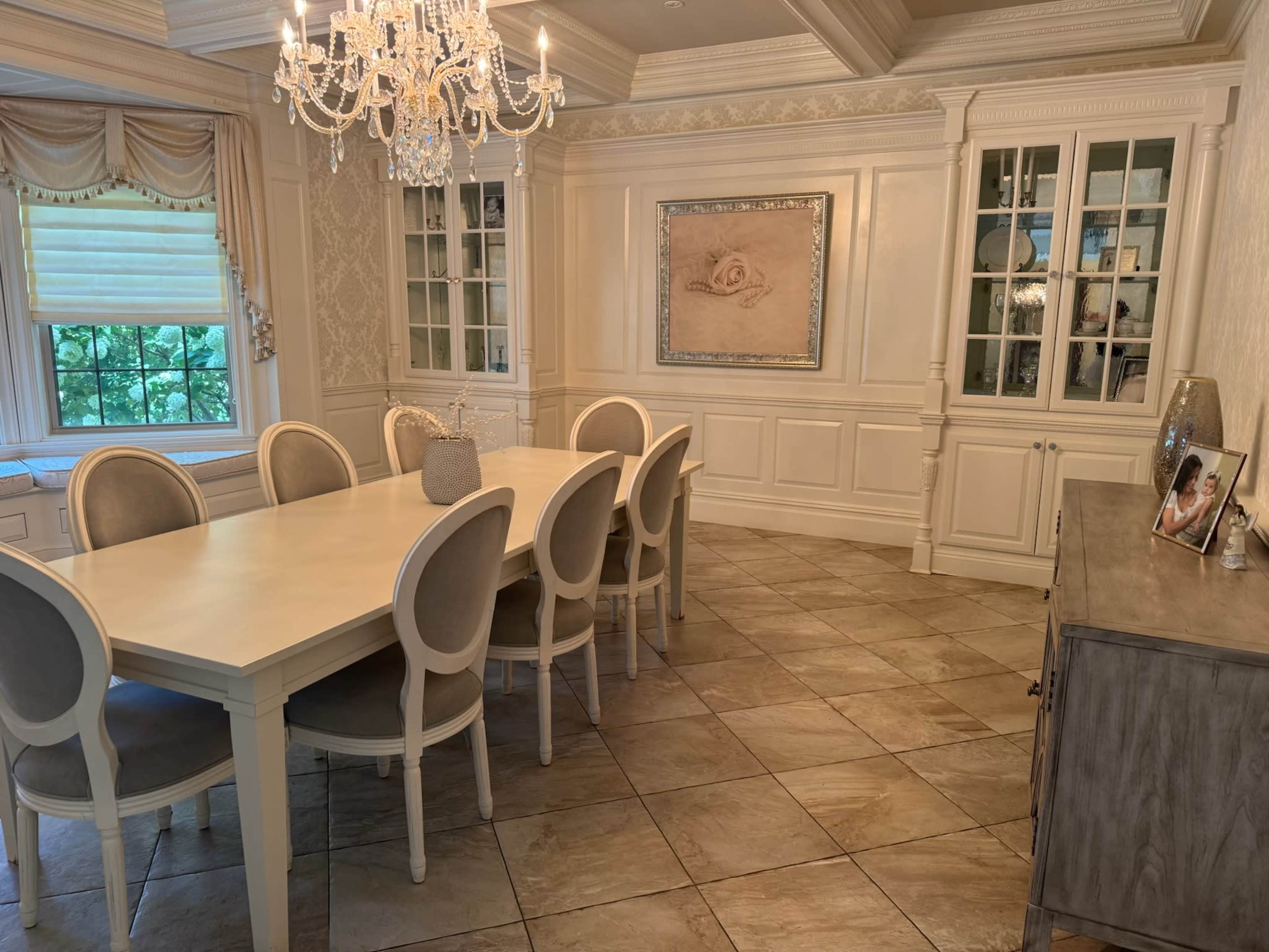 The image shows a well-lit dining room featuring a long white table surrounded by upholstered chairs, a chandelier overhead, and decorative cabinets along the walls.