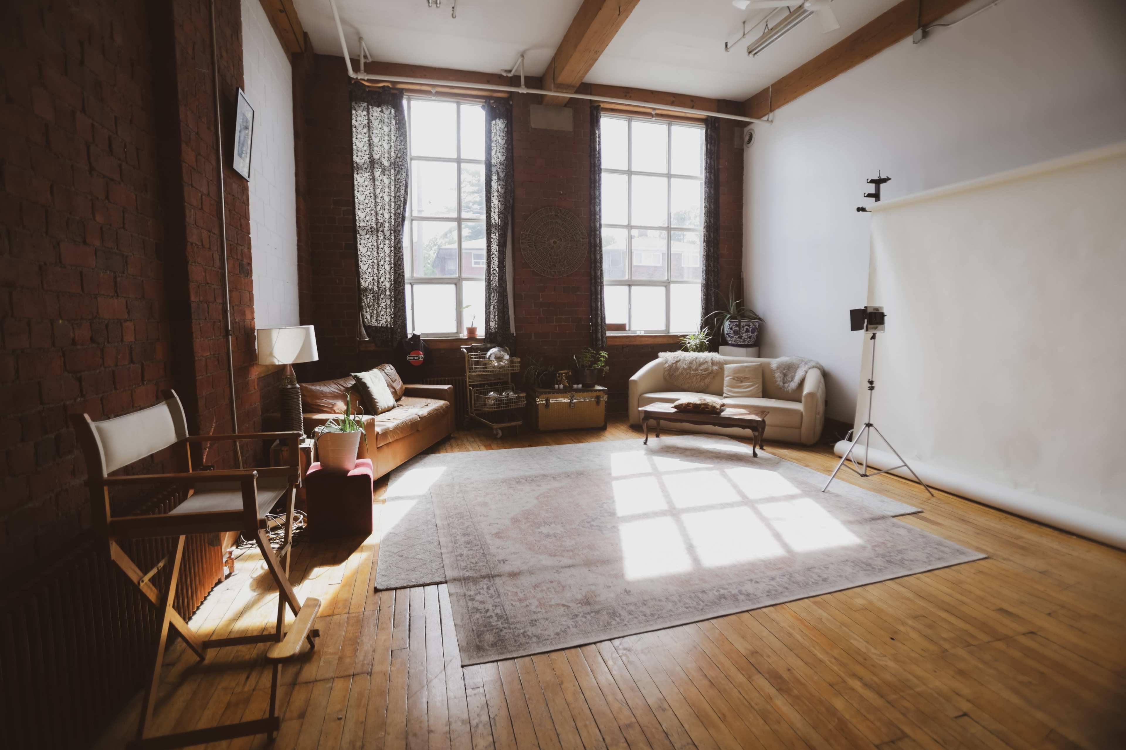 The image shows a spacious, well-lit photography studio with large windows, wooden floors, and a variety of seating arrangements, including a couch and chairs.