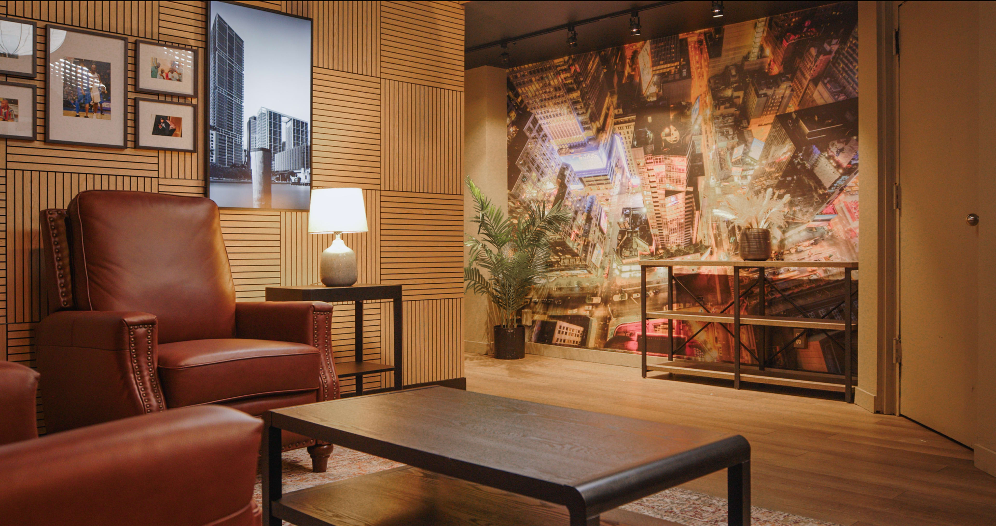 The image shows a cozy interior with brown leather chairs, a small coffee table, a lamp, and a wall decorated with a cityscape mural.